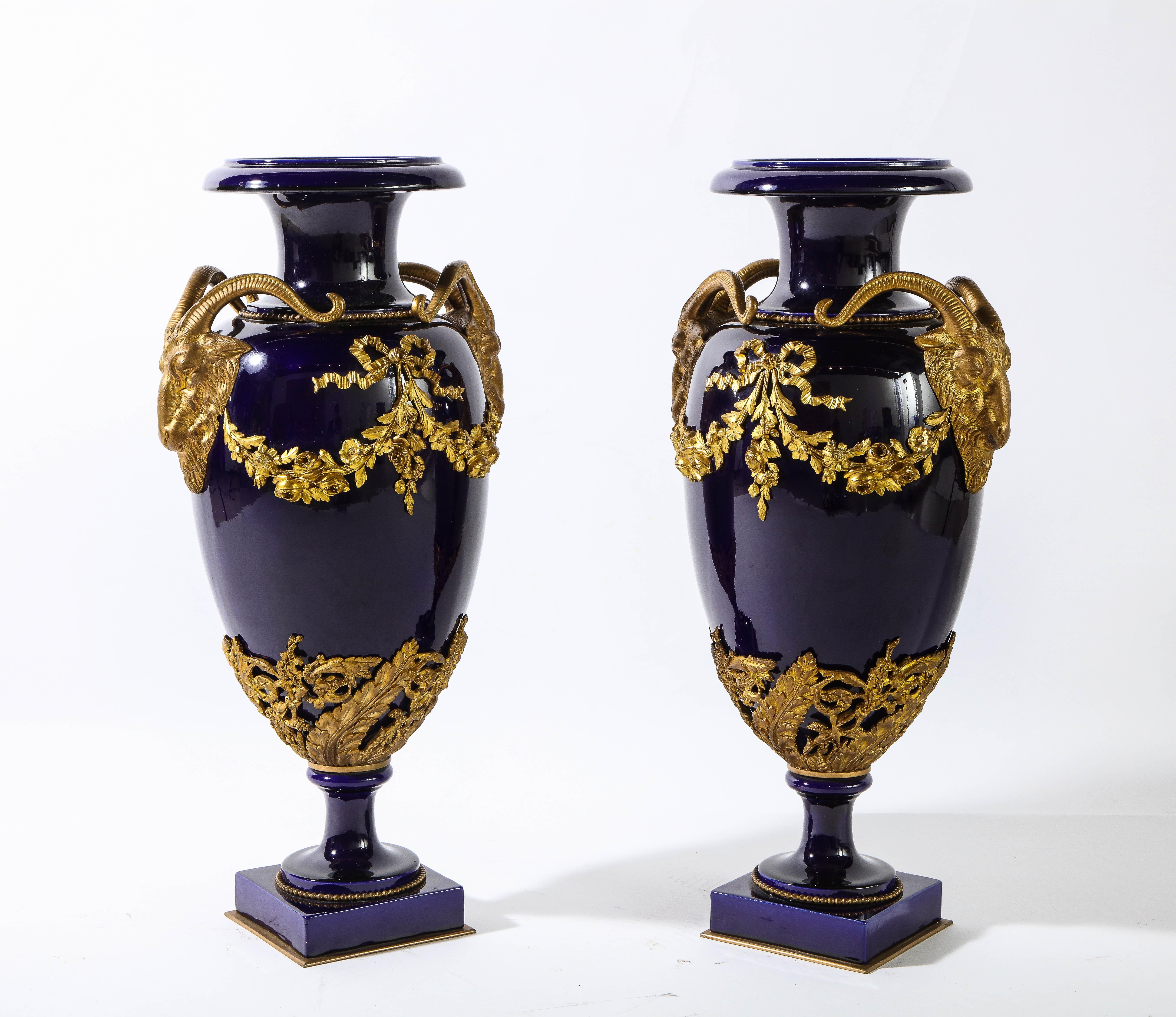 A fine pair of large antique French Louis XVI Sevres style cobalt blue porcelain and dore bronze mounted vases with wreaths, flowers, and ribbons, flanked on either end with a pair of rams head handles. This fabulous pair of porcelain vases have