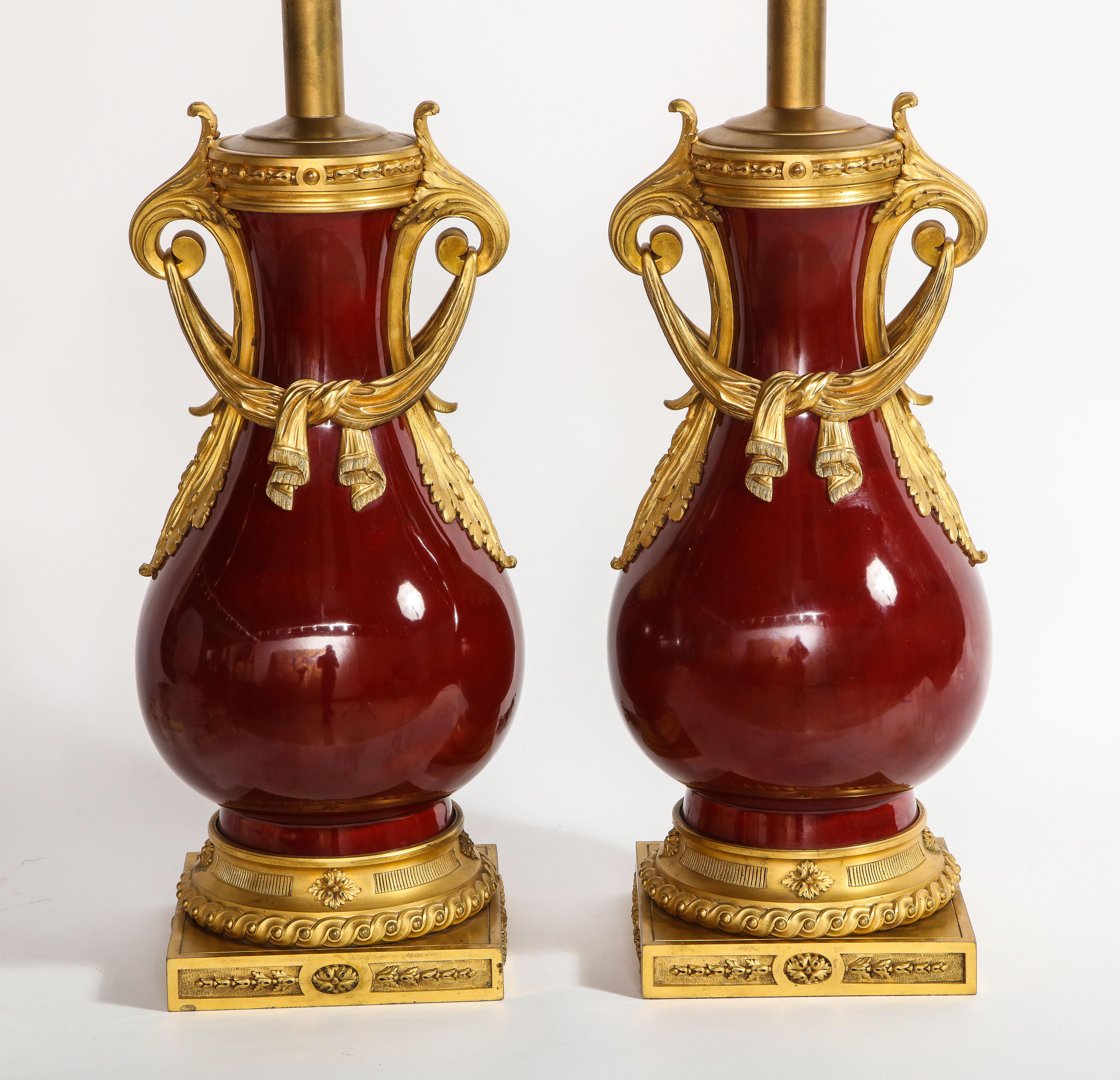 A very fine pair of antique French Louis XVI style dore bronze and Chinese sang de boeuf porcelain vases, later mounted as lamps, attributed to Alfred Beurdeley. Each baluster form mounted with twin gilt leafy cast handles, suspending gilt drapery
