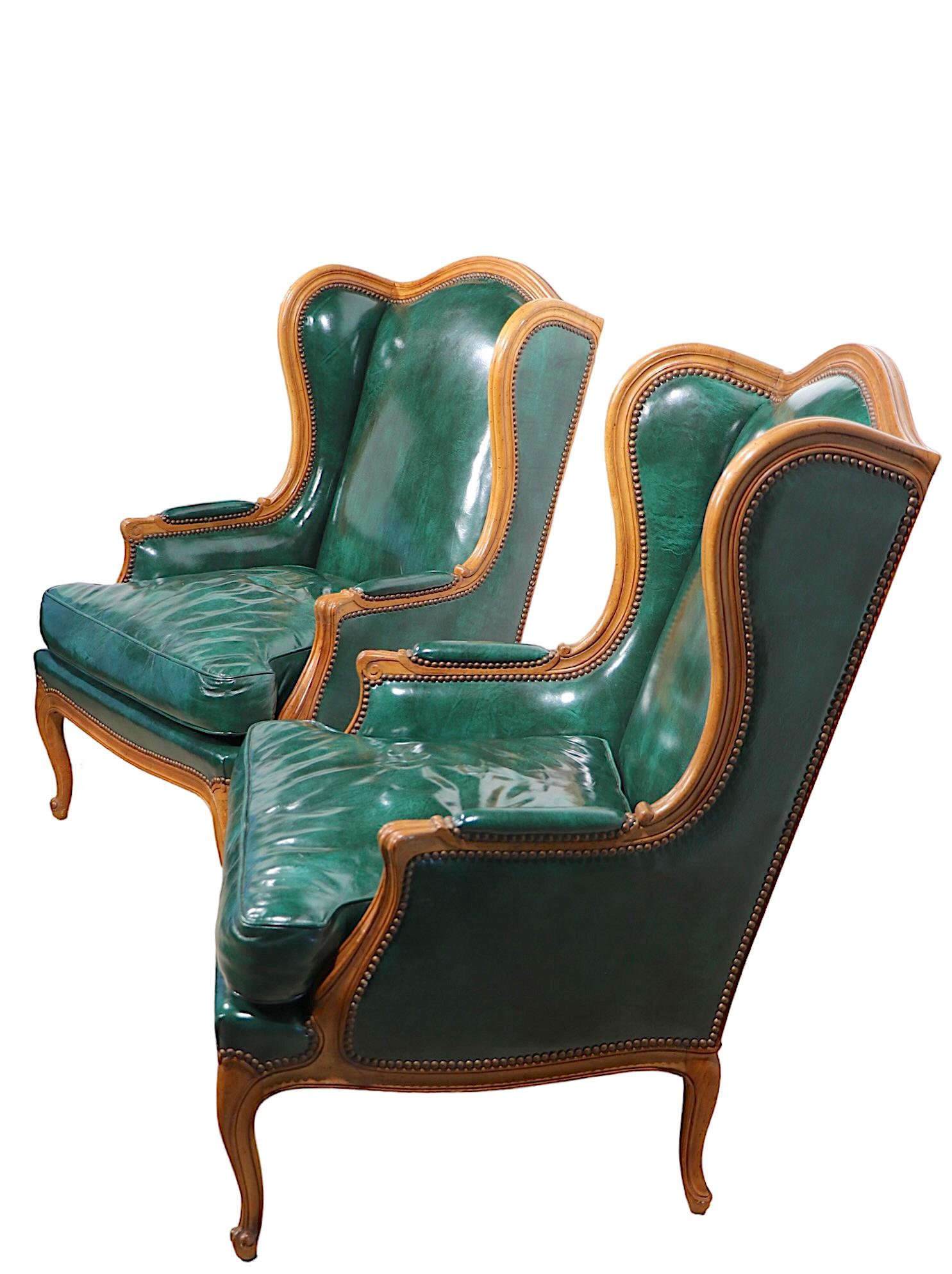 Voguish pair of wing back chairs, in the French Provincial style, by noted American furniture maker, Baker Furniture. The chairs feature leather upholstery with is trimmed with nailhead studs, on a hand carved frame of solid wood. Both chairs are in