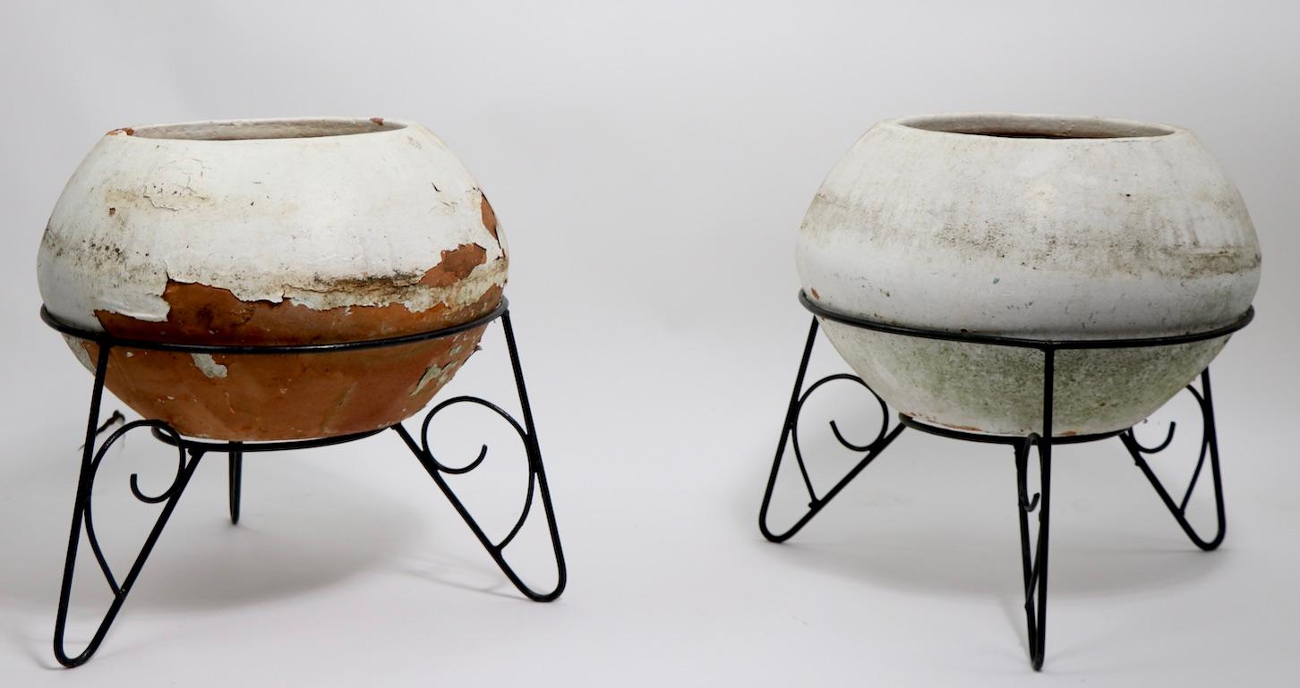 Pair of mid century planters in wrought iron stands. Planters rest in wrought iron base stands. Each ceramic planter is in later white paint finish, which is peeling away from the terracotta ground body. Planters show general cosmetic wear, normal