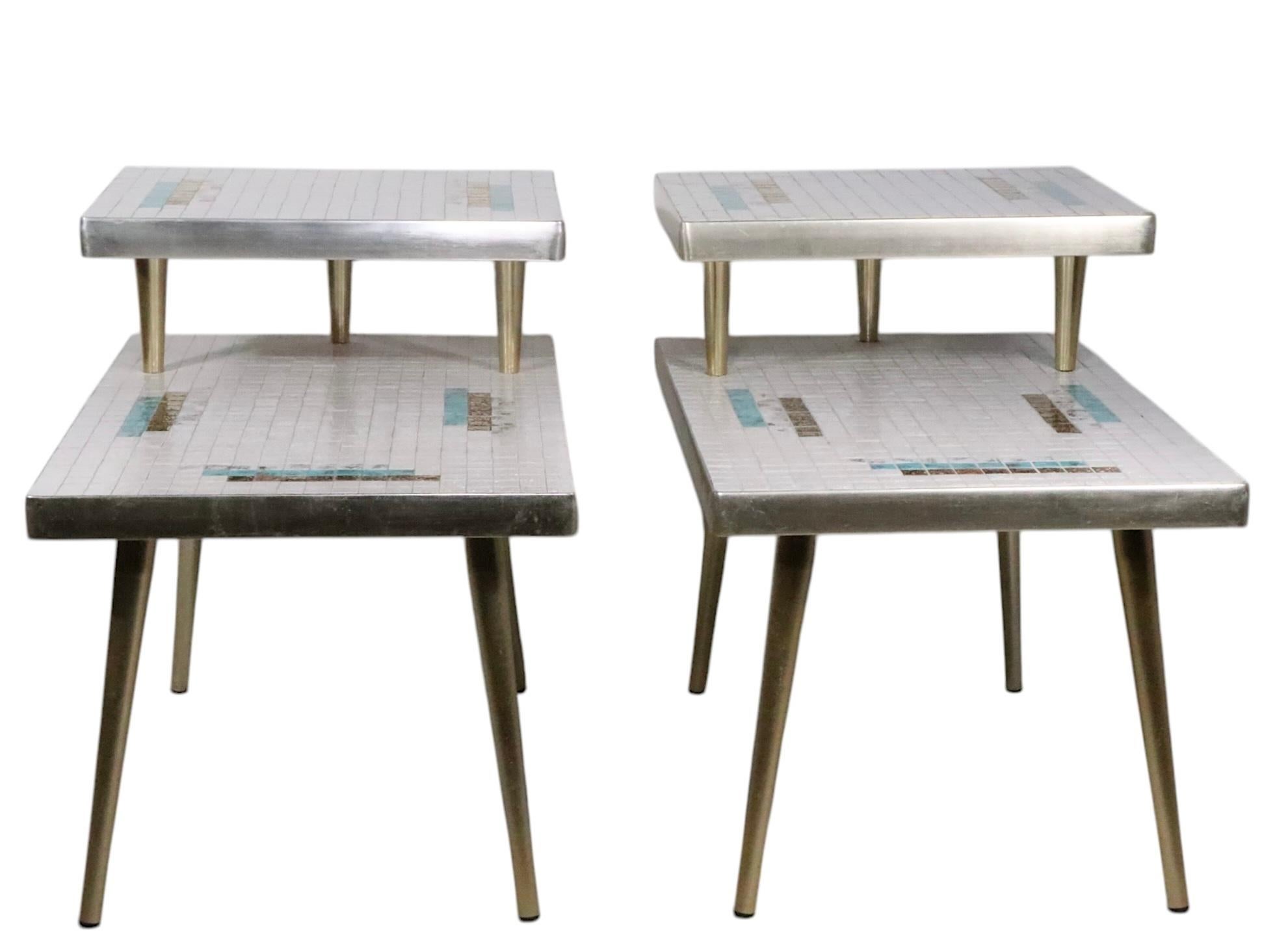 Pair of mid century step end, side tables having geometric ceramic tile tops, with anodized aluminum trim and tapered pole legs. The tables feature a square  upper shelf, which floats over the lower rectangular tile top. The ceramic tiles are in off