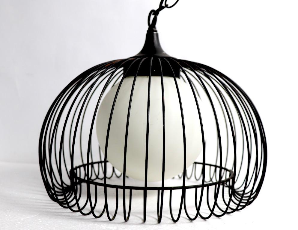 Una coppia di apparecchi a sospensione eleganti e raffinati, ognuno dei quali ha una struttura in ferro simile a una gabbia per uccelli che circonda un globo bianco smerigliato. Entrambe sono state originariamente progettate come luci swag, quindi