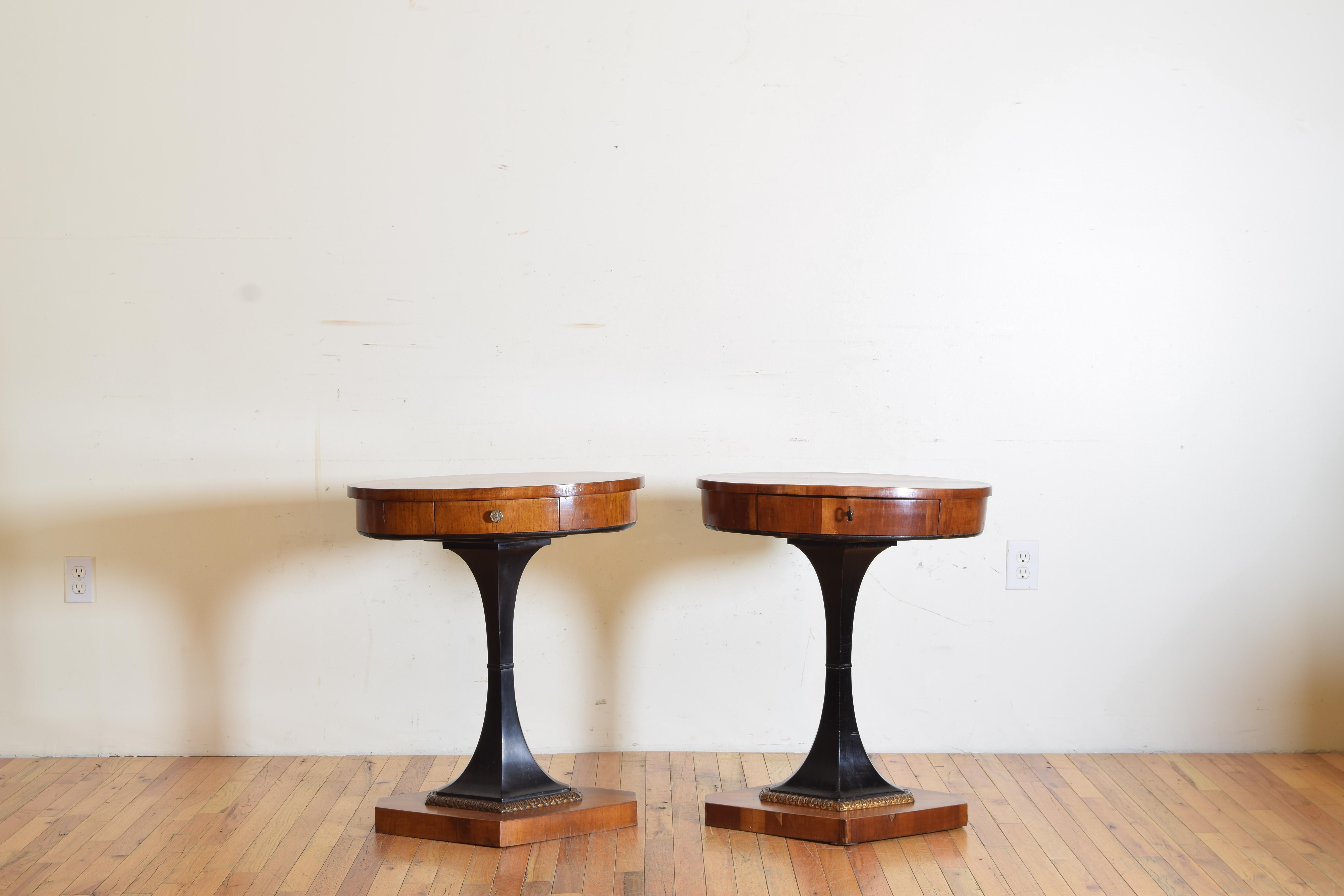 a NEAR pair, each of oval form and having one drawer with a brass pull, raised on shaped ebonized bases with carved gilt trim atop lozenge form bases, one of the tables from the early 19th century made of walnut, the newer table from the early 20th
