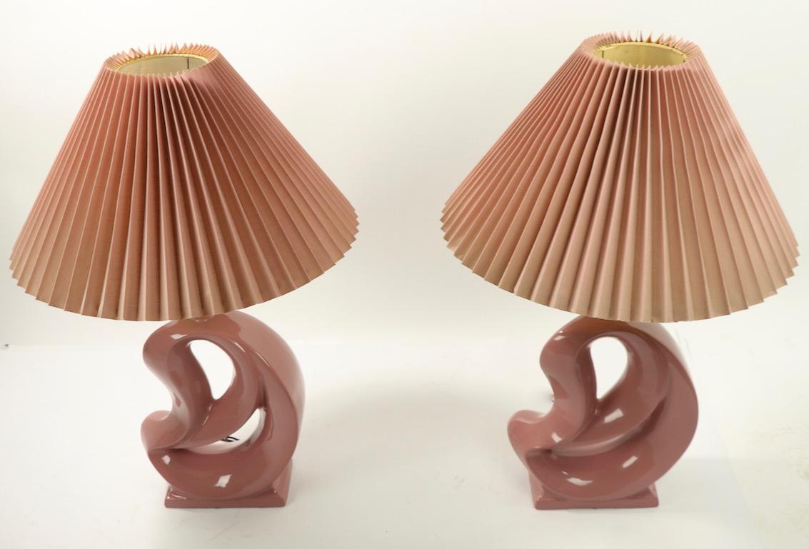 Pair of totally 1980s ceramic table lamps in high gloss mauve glaze finish, with the original conical pleated shades ( shades show dust and wear normal and consistent with age ). Both lamps accept standard size screw in bulbs and are in good working