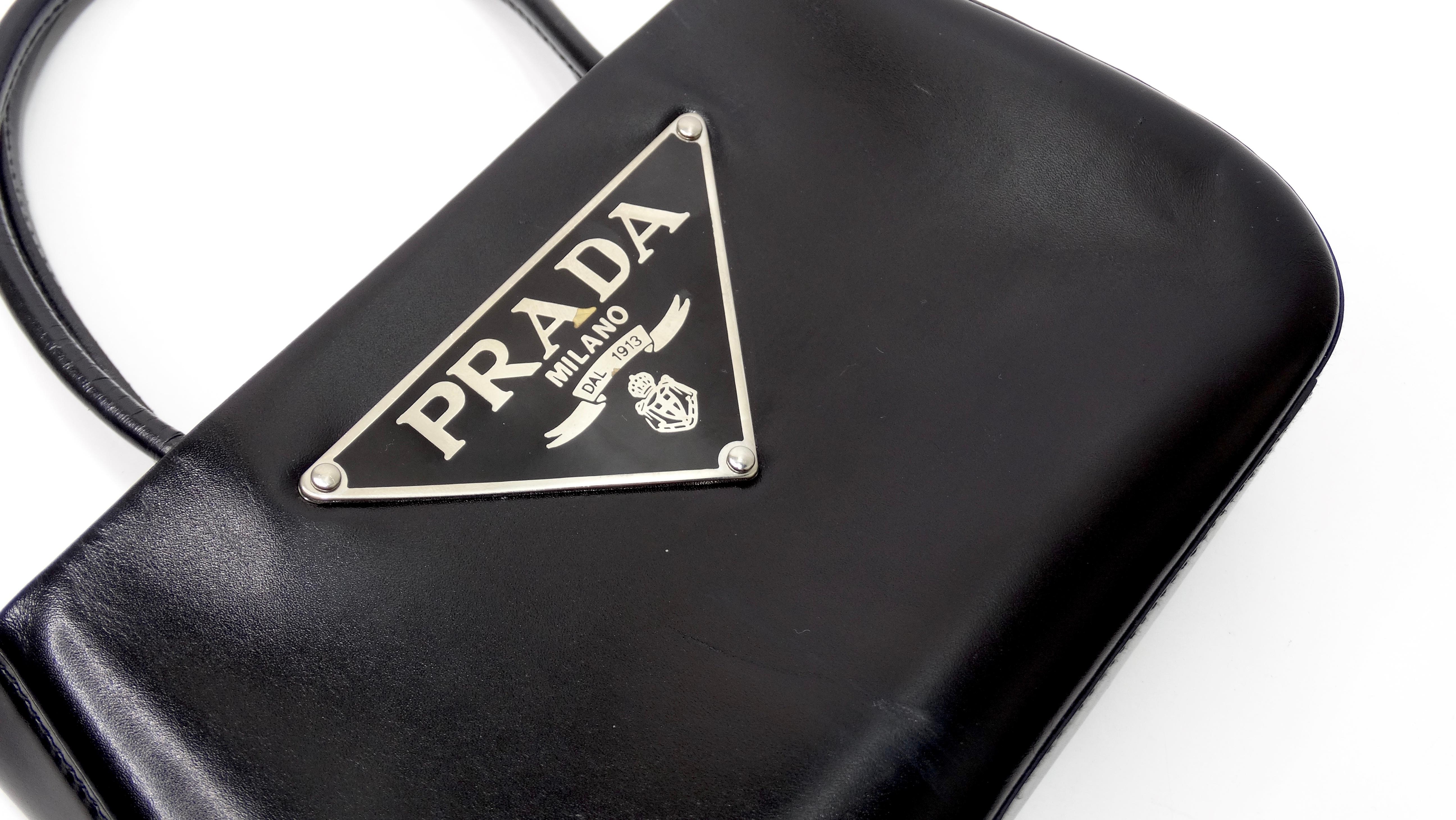 Add this cutie to your next look for a night out! Circa 1990s, this adorable mini handbag is crafted from smooth black calf skin leather with dual rolled top handles and the signature Prada emblem on the front face. Snap closure opens to a black