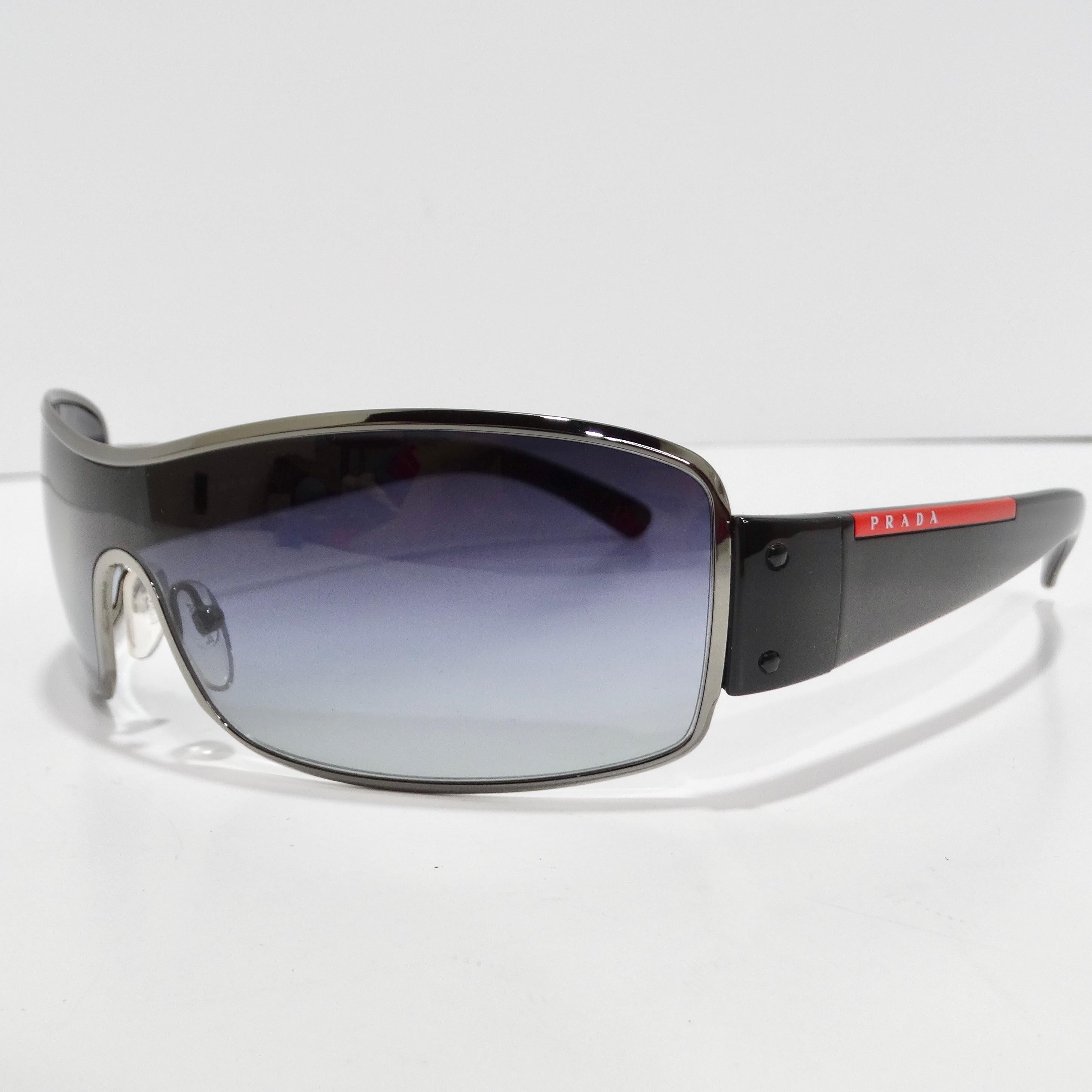 Step into the epitome of 1990s style with the Prada 1990s Silver Tone Shield Sunglasses. These classic shield-style sunglasses boast thin silver-tone rims, sleek black arms adorned with a red Prada logo, and stylish blue gradient lenses. The