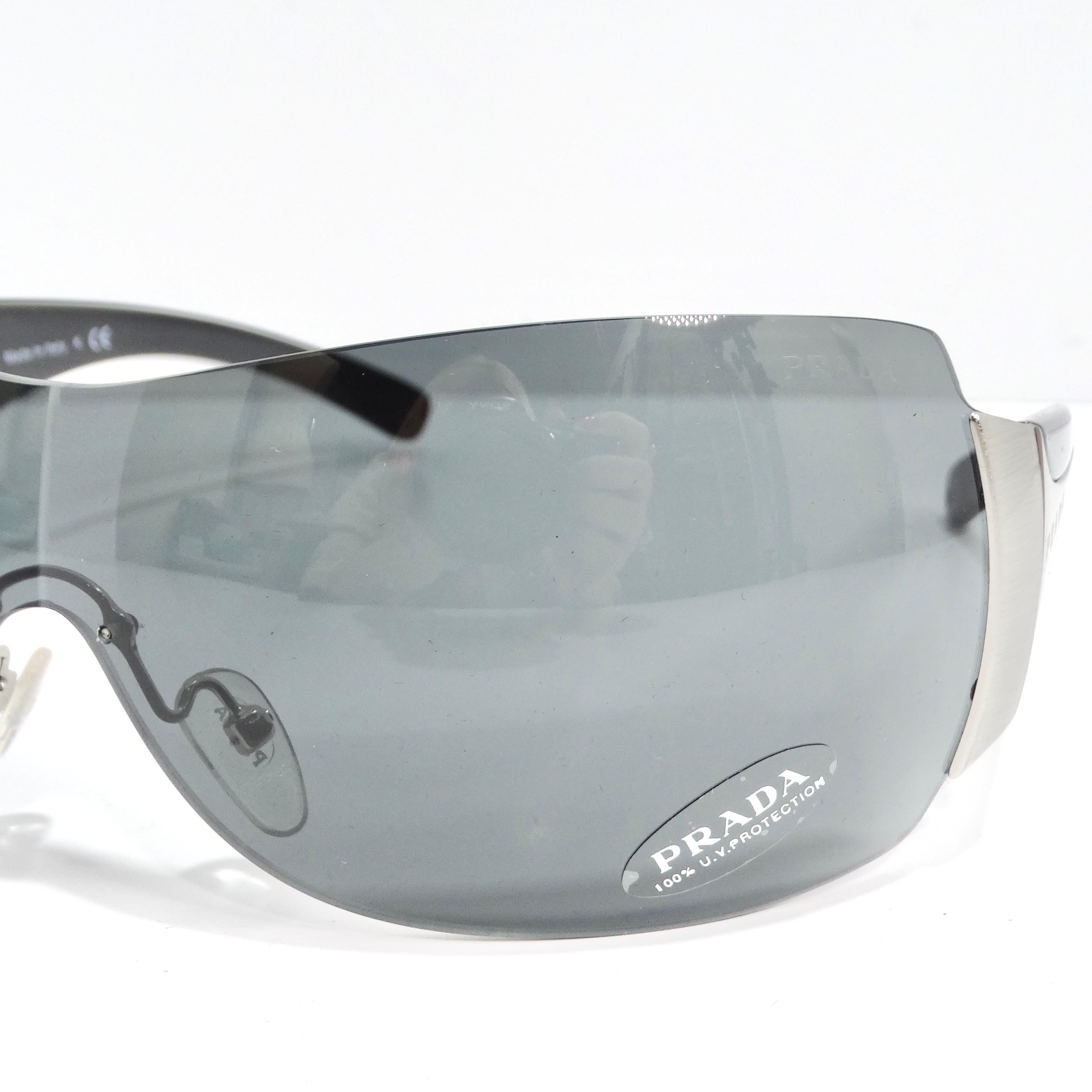 Step into the epitome of 1990s chic with the Prada 1990s Silver Tone Shield Sunglasses. These classic shield-style sunglasses boast thin silver-tone rims, sleek black arms adorned with silver-tone Prada logos, and stylish blue-grey lenses. The