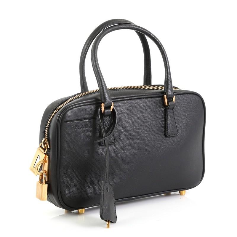 This Prada Bauletto Bag Saffiano Leather Small, crafted from black saffiano leather, features dual-rolled tall handles, raised Prada Milano logo, and gold-tone hardware. Its top zip closure opens to a black fabric interior with side zip pocket