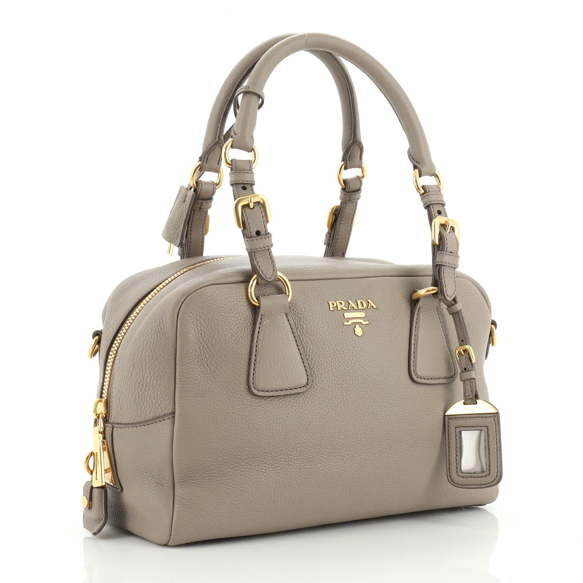 This Prada Bauletto Bag Vitello Daino Medium, crafted in neutral vitello daino leather, features dual top leather handles, Prada logo at the center and gold-tone hardware. Its zip closure opens to a gray fabric interior with side zip and slip