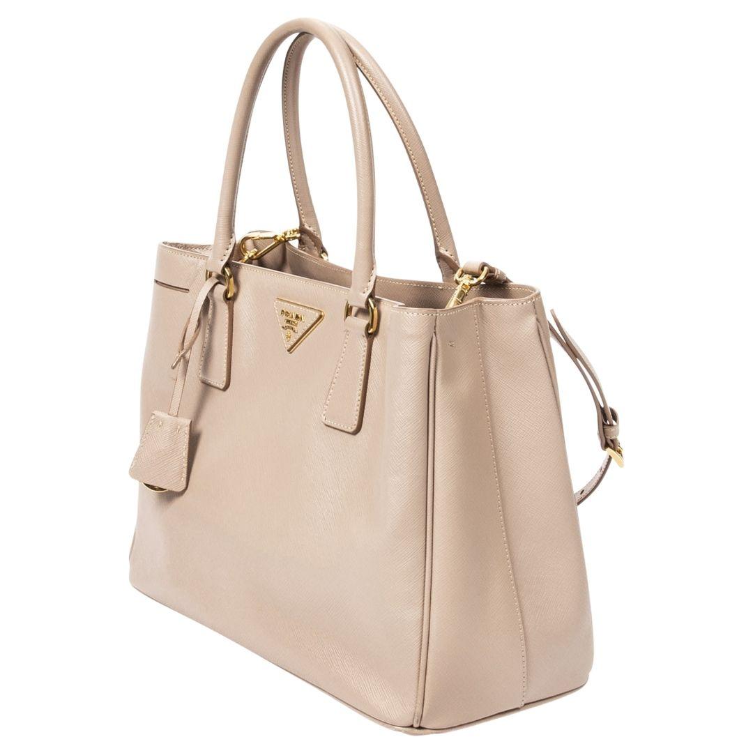 Elegantly crafted in beige Saffiano leather, this Prada tote features gold-tone hardware and a magnetic snap closure. The interior is divided into two compartments, with two zippered pockets and one slip pocket, all lined in logo
