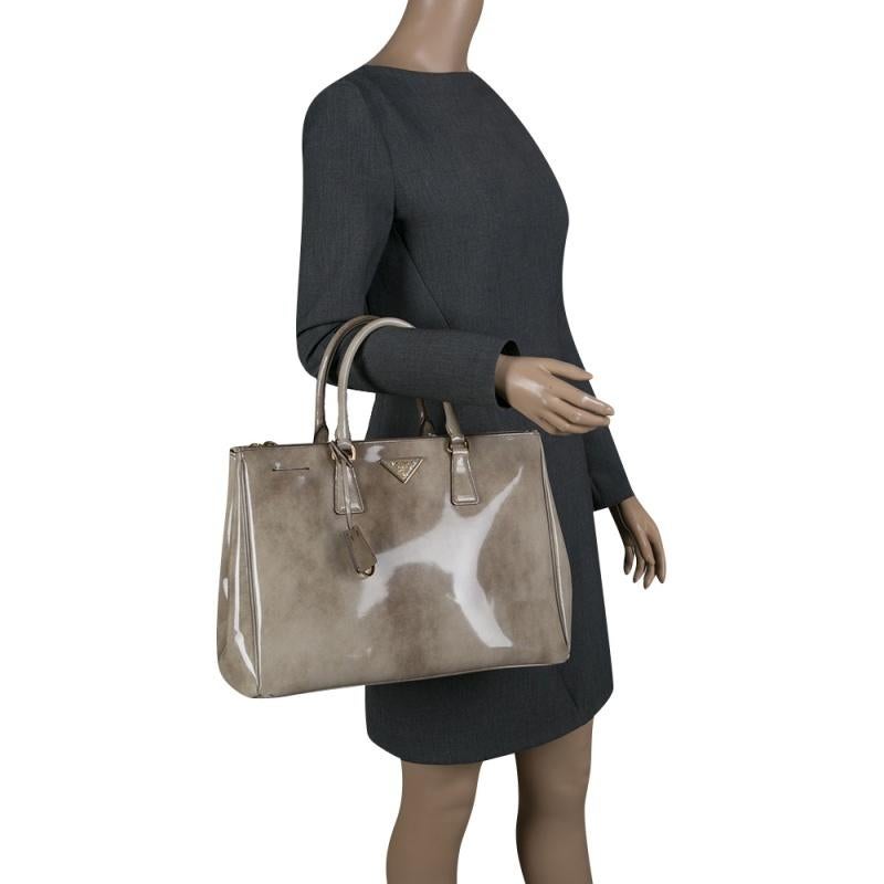 This elegant tote from Prada is crafted from patent leather and is perfect for daily use. The stunning bag features dual rolled top handles, a leather covered gold key ring and protective metal feet. The beige colored double zip tote has a nylon