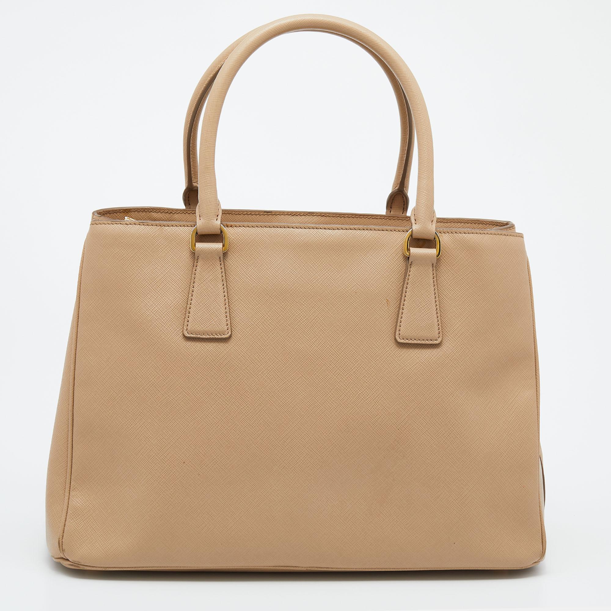 Loved for its classic appeal and functional design, Galleria is one of the most iconic bags from the house of Prada. This beauty in beige is crafted from Saffiano Lux leather and is equipped with two top handles, the triangular brand logo at the