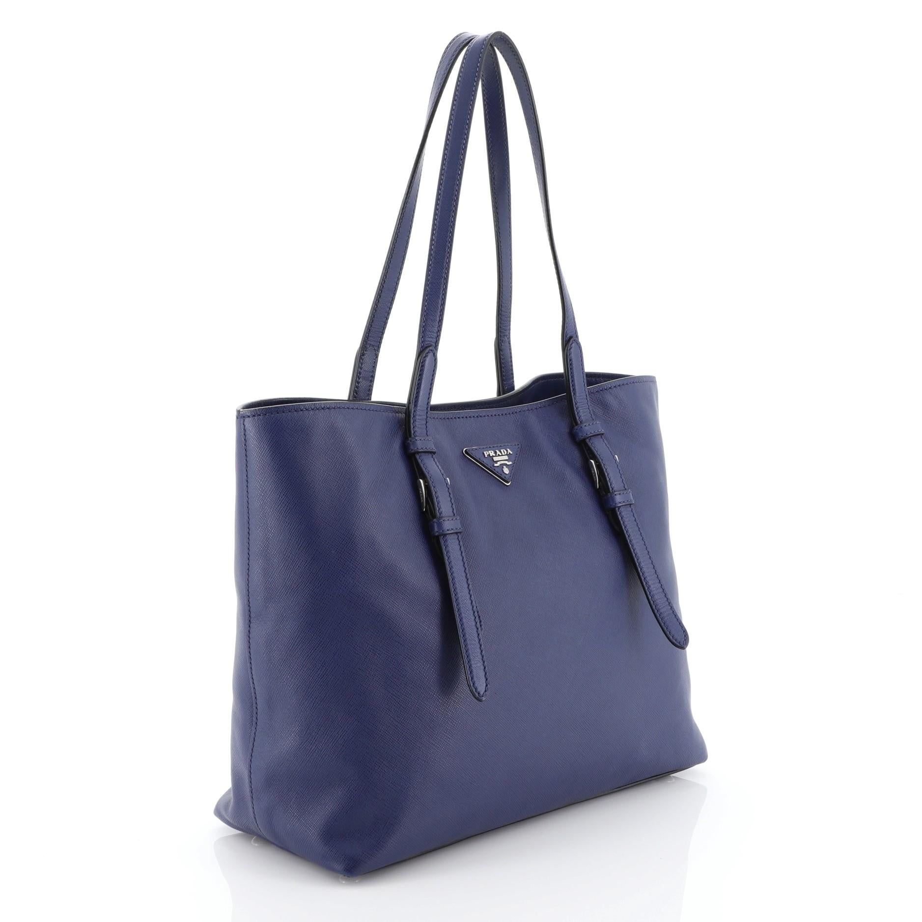 This Prada Belted Soft Tote Saffiano Leather Medium, crafted in blue saffiano leather, features dual flat leather handles with belted accent, protective base studs, triangle logo at the center and silver-tone hardware. Its magnetic snap closure