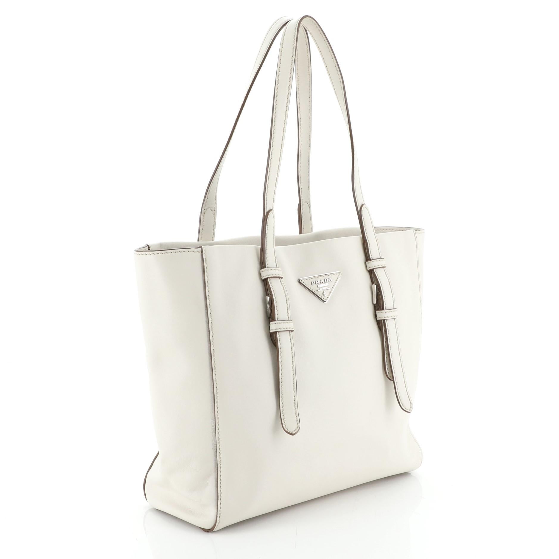 This Prada Belted Tote Soft Calfskin Medium, crafted from white soft calfskin leather, features dual tall flat handles with belt details, raised Prada logo, and silver-tone hardware. Its zip closure opens to a brown fabric interior with zip and slip