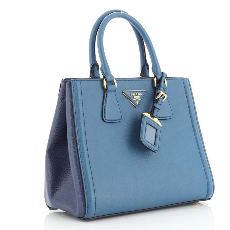 This Prada Bicolor Lux Convertible Open Tote Saffiano Leather Small, crafted in blue saffiano leather, features dual rolled leather handles, side snap buttons, protective base studs, and gold-tone hardware. Its snap button closure opens to a blue