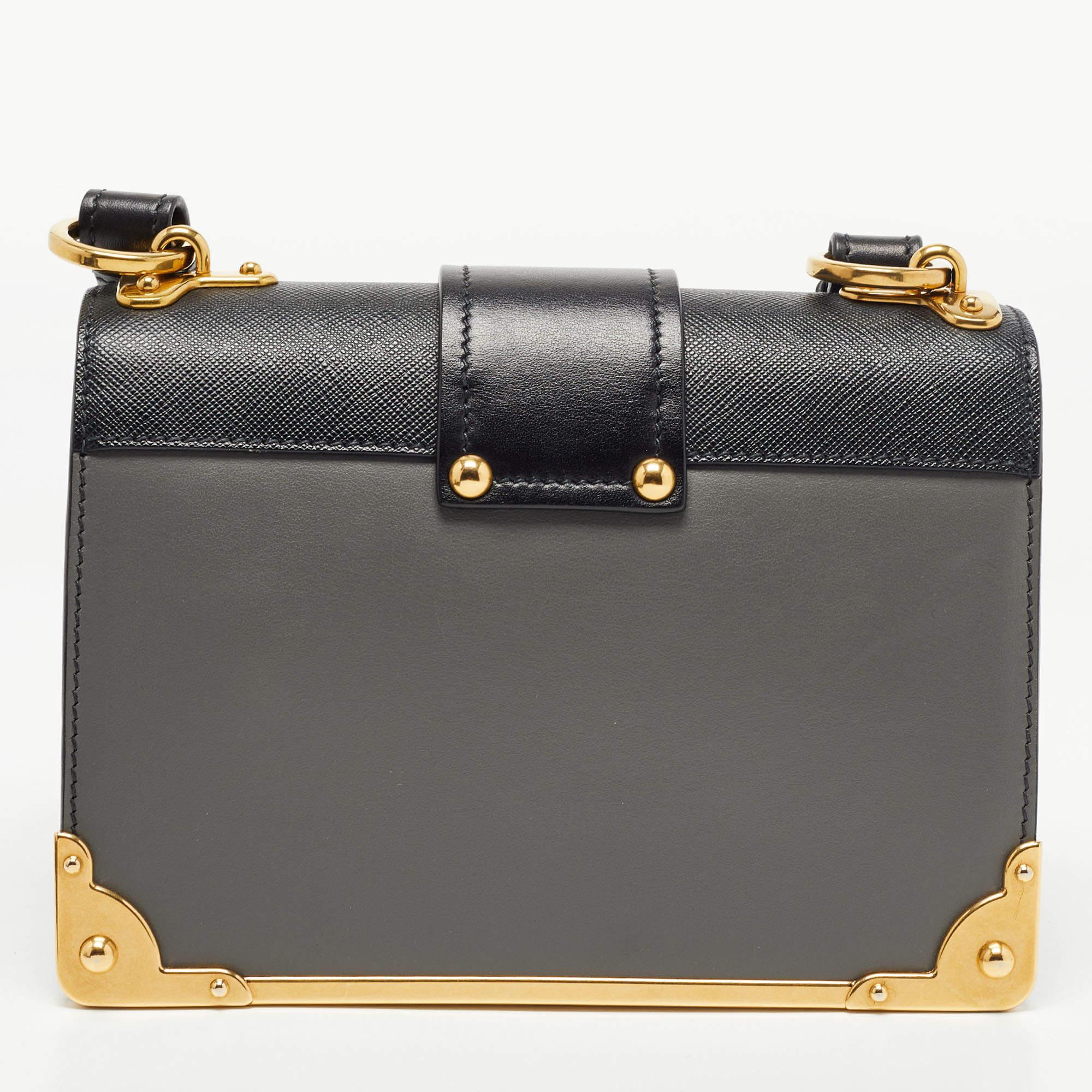 Inspired by valuable books from ancient times, the Cahier by Prada is a best-seller. This shoulder bag is crafted in Italy with black and grey leather. It features gold-tone trims that add a touch of contrast. The strap closure with the brand logo
