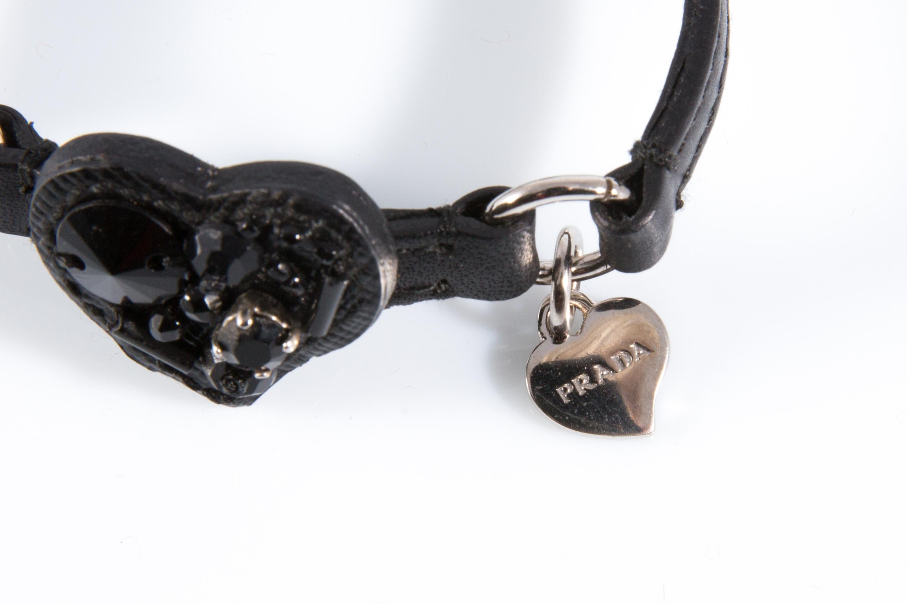 Bracelet cœur en cuir noir Prada avec forme de cœur embellie, bracelet en cuir noir, métal argenté, breloque cœur logo. 
Circa 2000s
En bon état vintage. Fait en Italie. 
Longueur maximale : 8,6 pouces (22 cm)
Largeur : 2,5 cm (1 pouce)
Nous