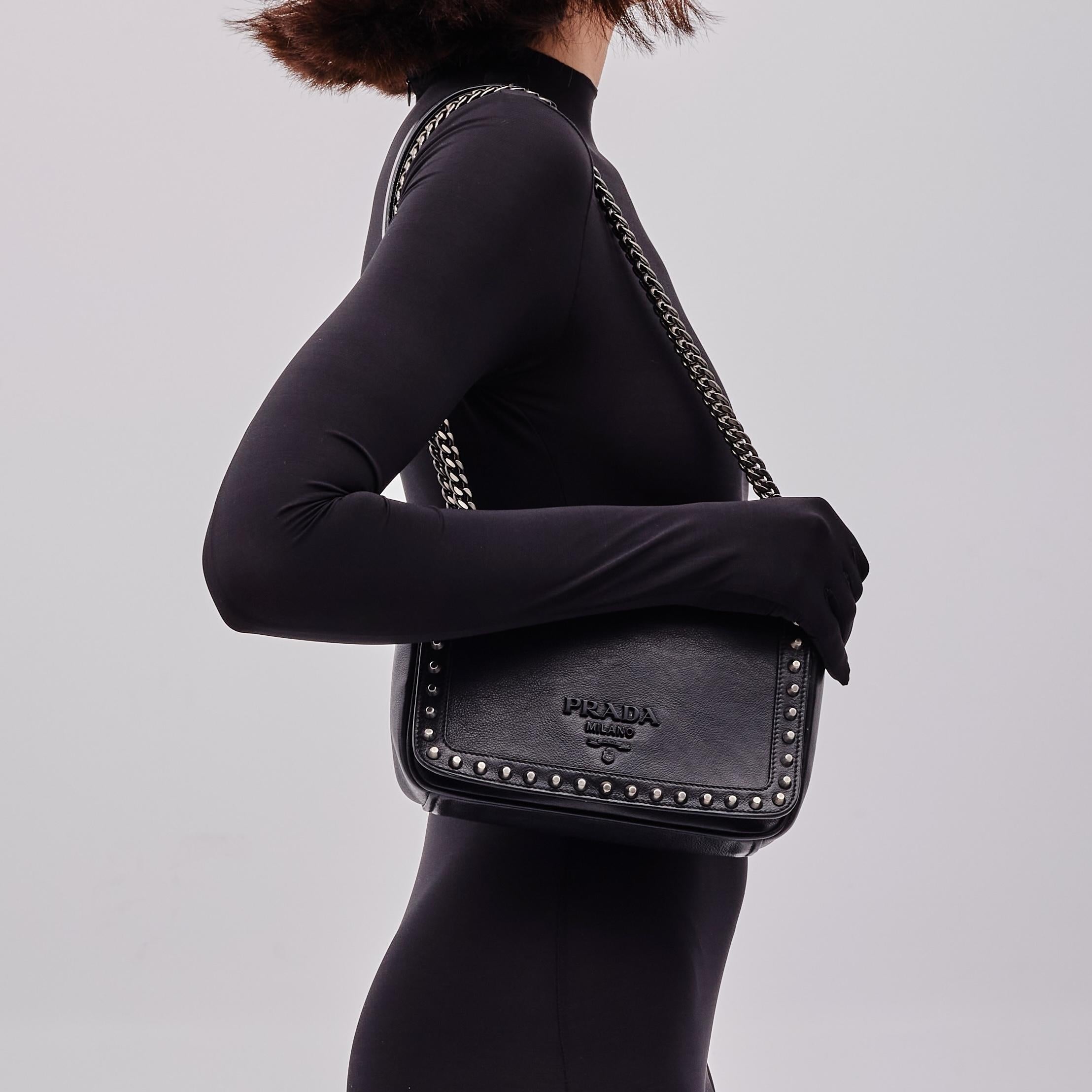 Este elegante bolso bandolera está fabricado en piel de becerro granulada de color negro. Este elegante bolso cruzado tiene una larga correa de cadena de rutenio / plata oscura con una hombrera de piel negra, tachuelas de plata oscura y un llamativo