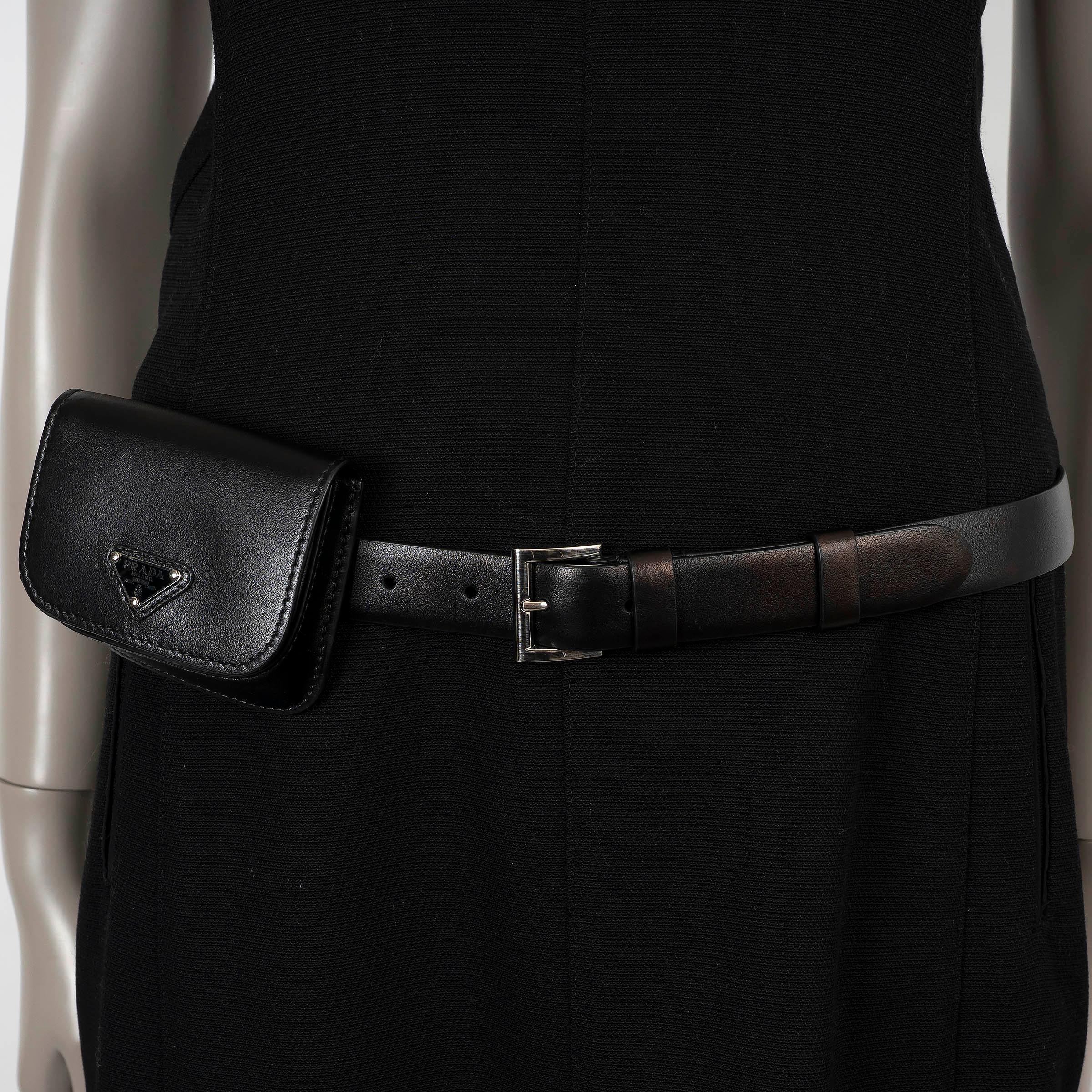 Questa mini borsa con cintura di Prada, autentica al 100%, è realizzata in pelle nera. È dotata di un'elegante patta con il caratteristico logo a triangolo smaltato. La borsa si chiude saldamente con una chiusura magnetica ed è foderata in nylon