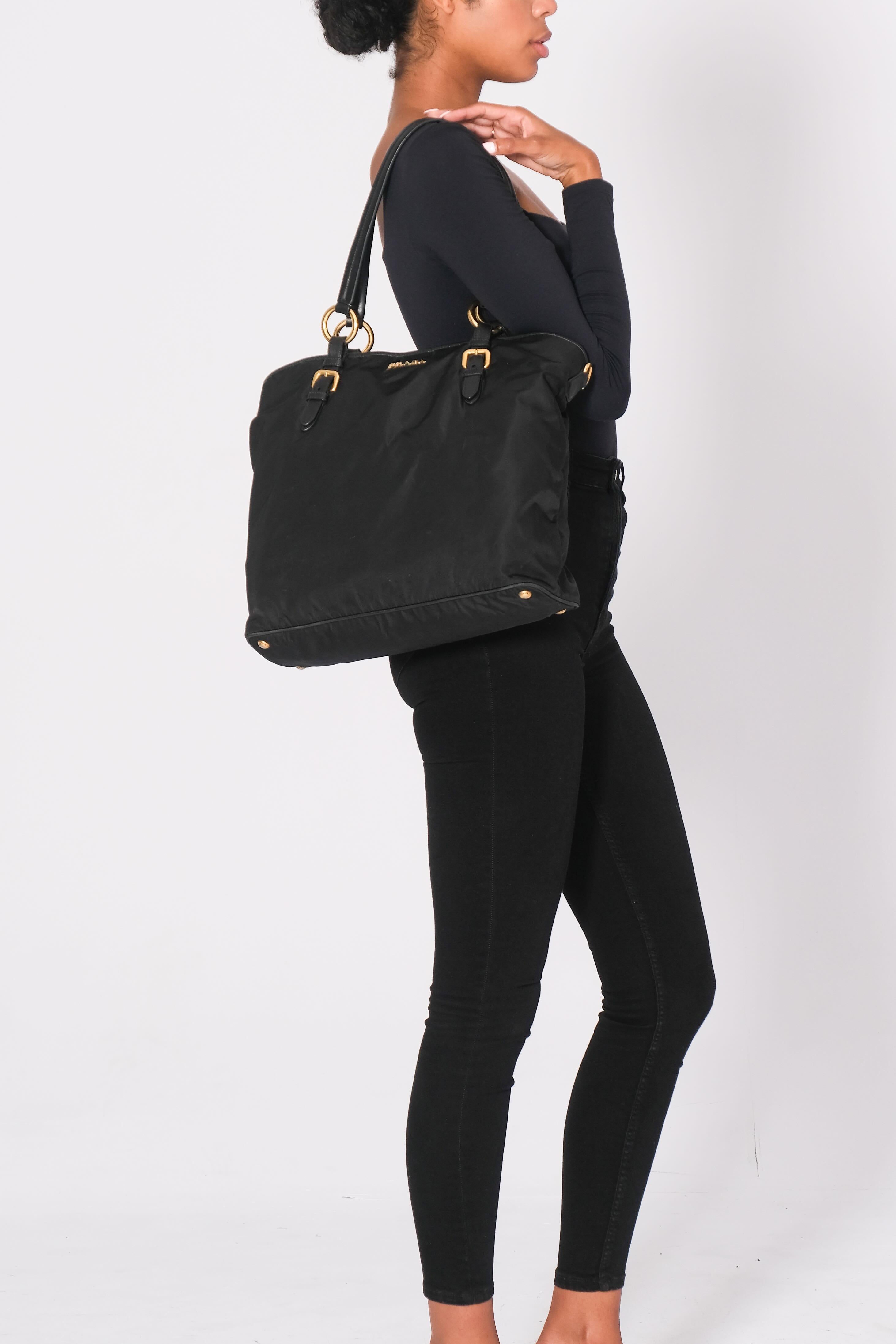 This Prada tote is constructed of Nero Vitello Daino nylon with gold-tone hardware and leather trim. It features dual shoulder straps, leather trim embellishments. snap closure at the top, a luggage tag, and has protective feet at the base. The