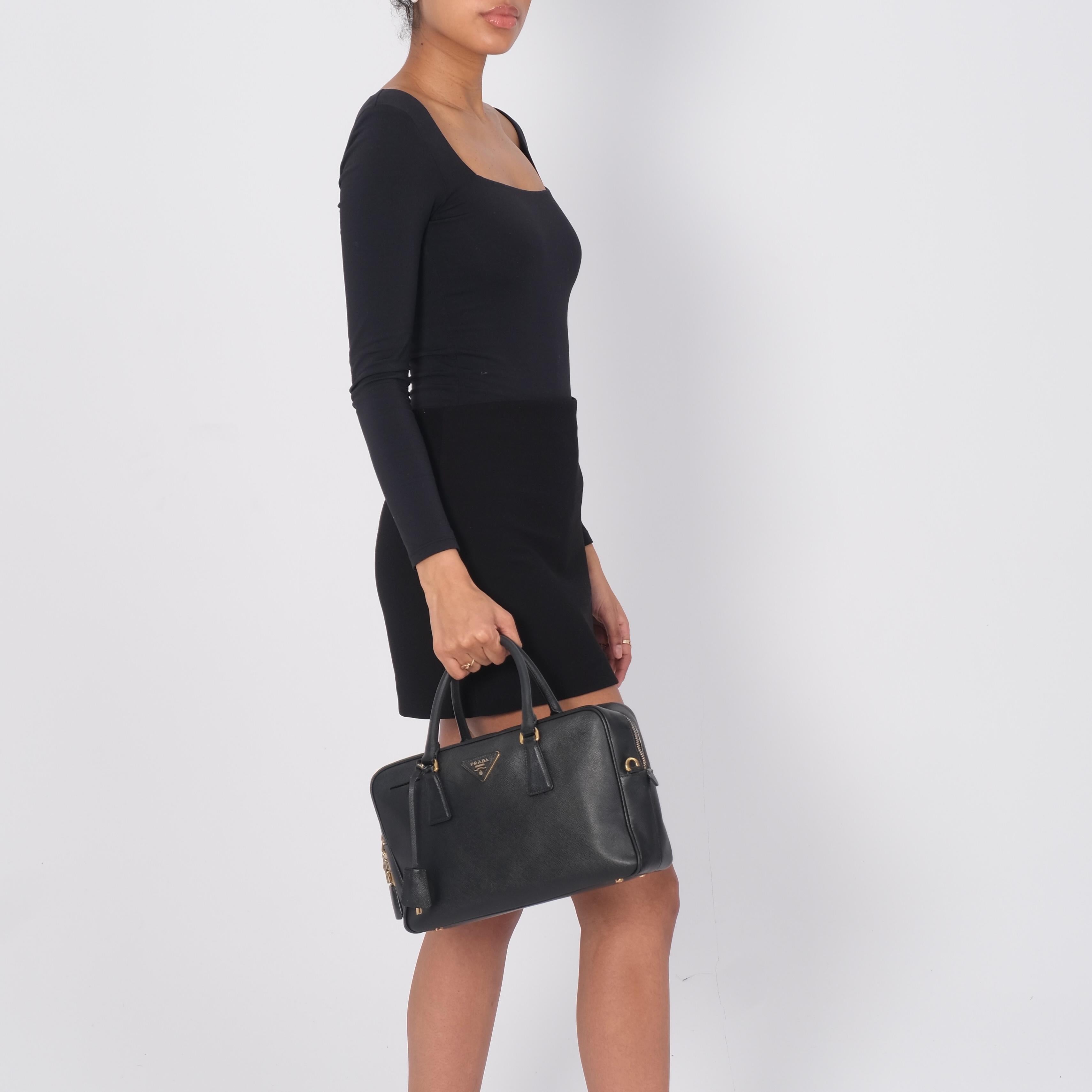 Te presentamos la bandolera Prada fabricada en piel saffiano lux negra. El bolso presenta herrajes dorados, dos asas superiores enrolladas de piel y pies protectores en la base. La cremallera superior se abre a un forro de jacquard con logotipo y un
