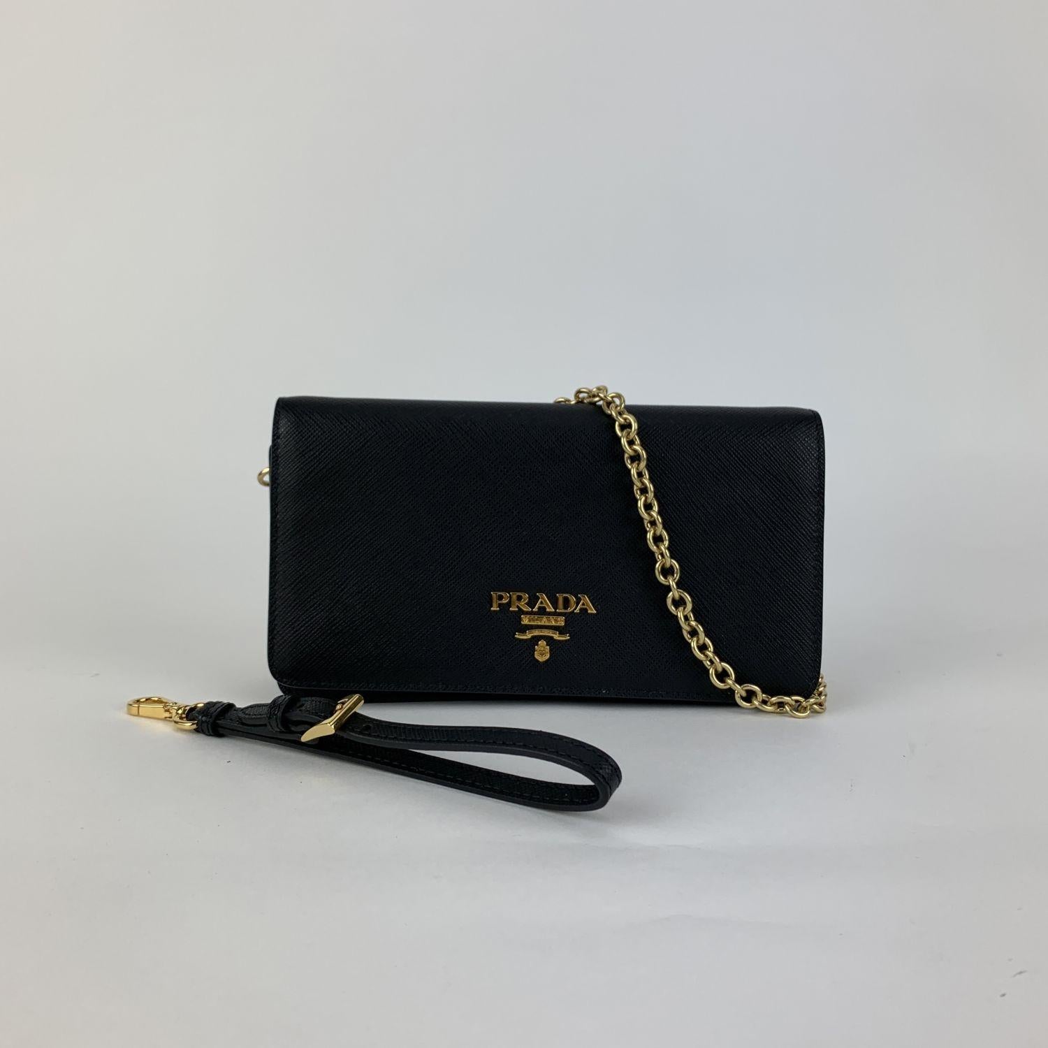 Prada black Saffiano leather flap continental wallet on chain/WOC, mod. 1DH029. Gold metal hardware. PRADA Metal lettering logo. Double magnetic button closure. 2 credit card slots. 1 coin compartment with zipper, 2 bill compartment and 3 open