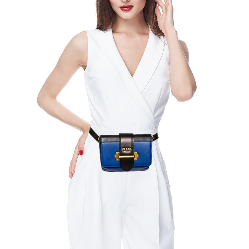 Inspired by valuable books from ancient times, the Cahier by Prada is a best-seller. This belt bag is crafted in Italy with blue and black leather. It features a gold-tone loop at the front that adds a touch of contrast. The strap closure with the
