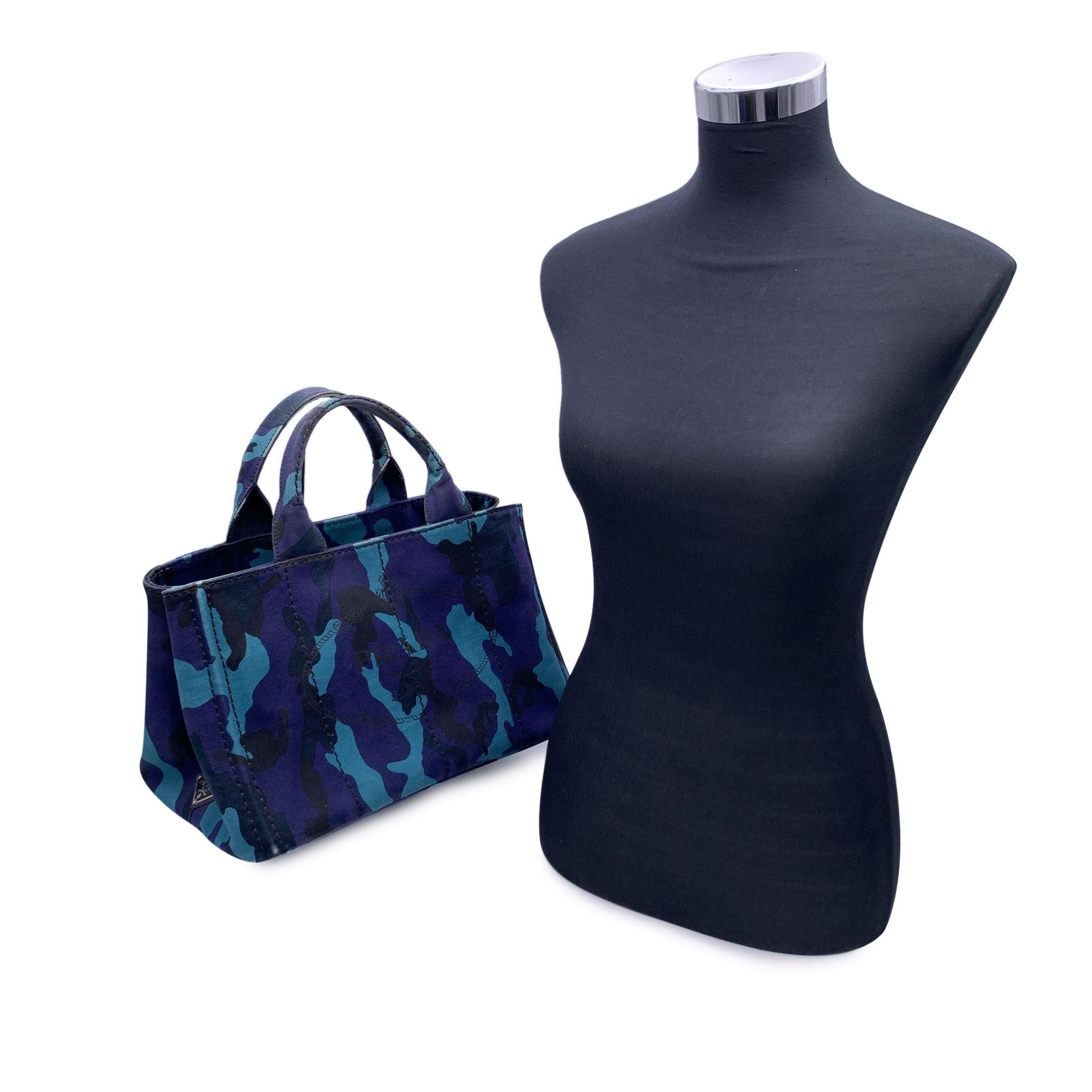 Prada Canapa tote bag in blue hemp Camouflage canvas with printed Prada logo and Prada triangle logo on the sides. Canvas handle. Open top with side snap buttons. Canvas interior with 2 side zip pockets and 3 open pockets and black hemp lining. 4