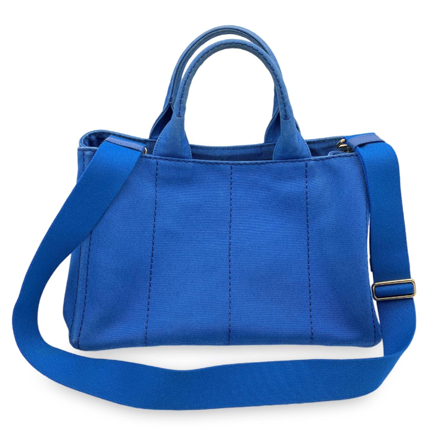 Prada large Canapa 2 way tote bag in blue hemp canvas with printed Prada logo and Prada triangle logo on the sides. Canvas handles. Removable canvas shoulder strap. Open top with side snap buttons. Canvas interior with 2 zip pockets and 3 open