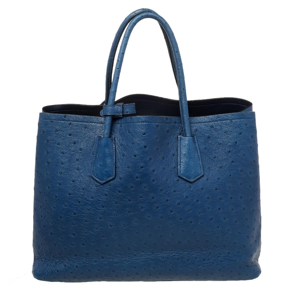 This magnificent Prada tote comes meticulously crafted from ostrich skin to add a royal touch to your closet. It has been shaped beautifully and equipped with two top handles for you to flaunt it. The blue piece is complete with protective metal