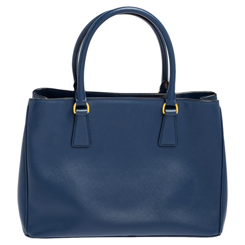 Amata per il suo fascino classico e il suo design funzionale, la Galleria è una delle borse più iconiche e popolari della casa di Prada. Questa bellezza in blu è realizzata in pelle Saffiano Lux ed è dotata di due manici superiori, logo del marchio