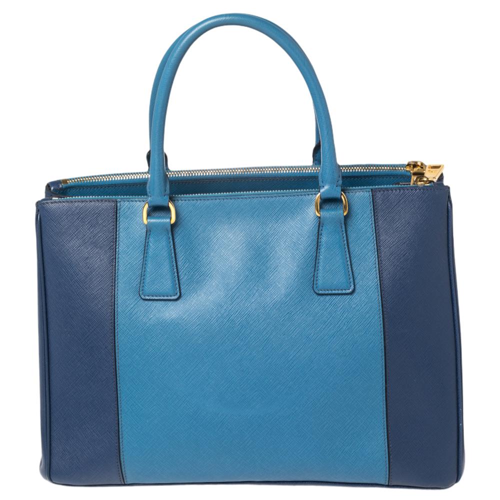 Loved for its classic appeal and functional design, Galleria is one of the most iconic and popular bags from the house of Prada. This beauty in bicolored blue is crafted from Saffiano Lux leather and is equipped with two top handles, the brand logo