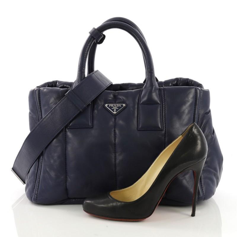 This Prada Bomber Convertible Tote Nappa Leather Medium, crafted in blue nappa leather, features dual rolled handles and silver-tone hardware. Its top zip closure opens to a black fabric interior with zip and slip pockets. **Note: Shoe photographed