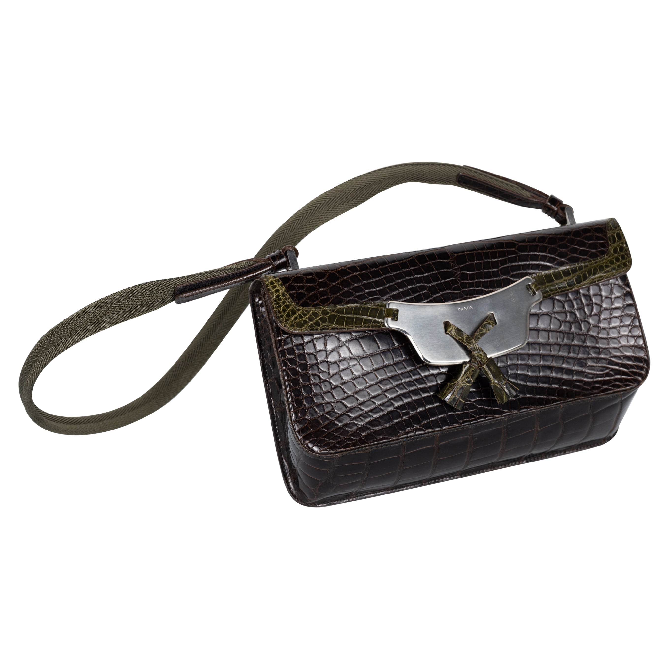 This Prada handbag features a luxurious crocodile leather exterior with a dark brown finish, showcasing a sophisticated and textured look. The flap closure is accented with an olive green trim and a striking silver-toned metal plate engraved with