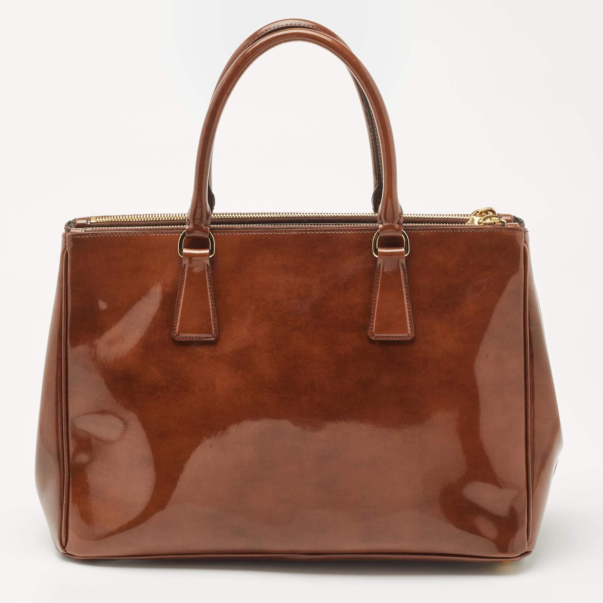 Feminine in shape and grand in design, this Double Zip tote by Prada will be a valuable addition to your closet. It has been crafted from patent leather and styled minimally with gold-tone hardware. It comes with dual handles at the top, two zip