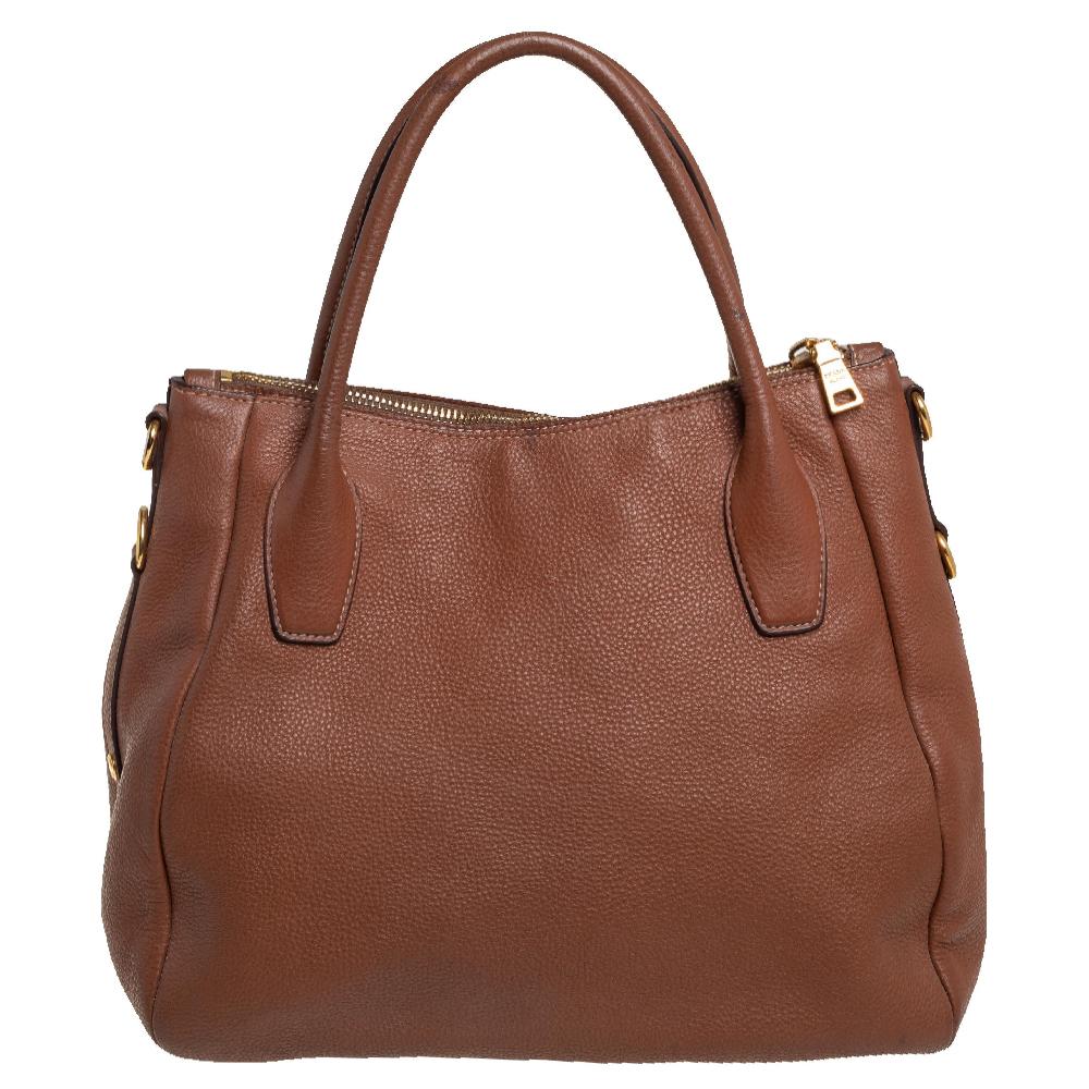 This Prada Sacca 2 Manici tote is made of Vitello Daino leather. It features rolled top handles and gold-tone hardware. A recognizable Prada logo sits at the front. The interior is lined with durable nylon and comprises a slip pocket and enough