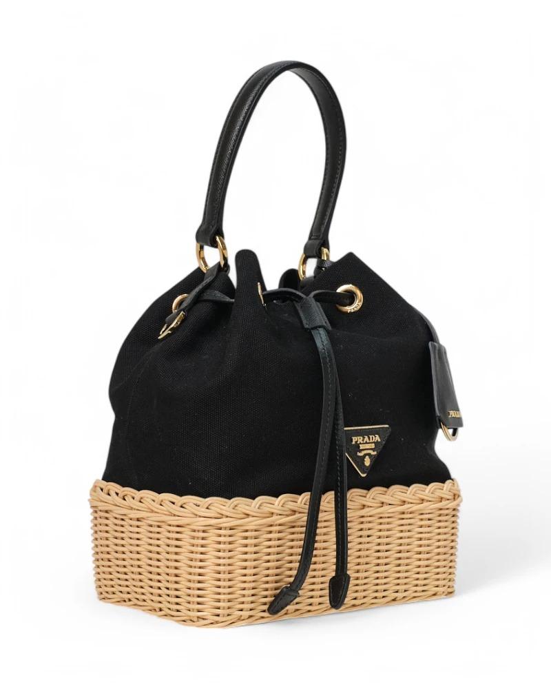 Prada signature bag, Bucket model in size small, made of black canvas with wicker trim and gold hardware. Featuring a drawstring closure, internally lined with logo fabric with a small pocket, the bag is quite roomy. Equipped with a handle and a