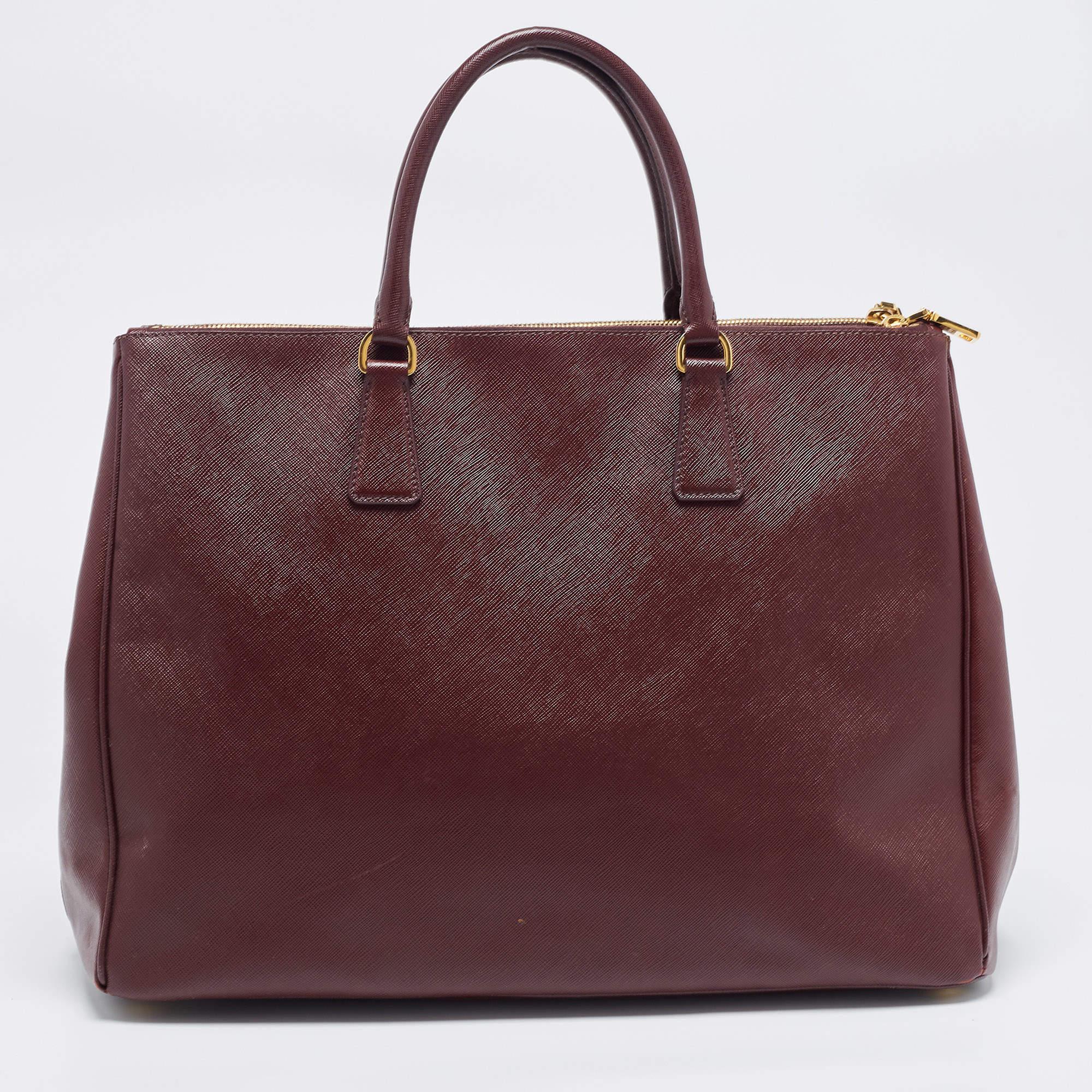 This extra-large Executive tote by Prada will be a loved addition to your closet. It has been crafted from Saffiano leather and styled with gold-tone hardware. The burgundy bag comes with two top handles, double zip compartments, and a spacious