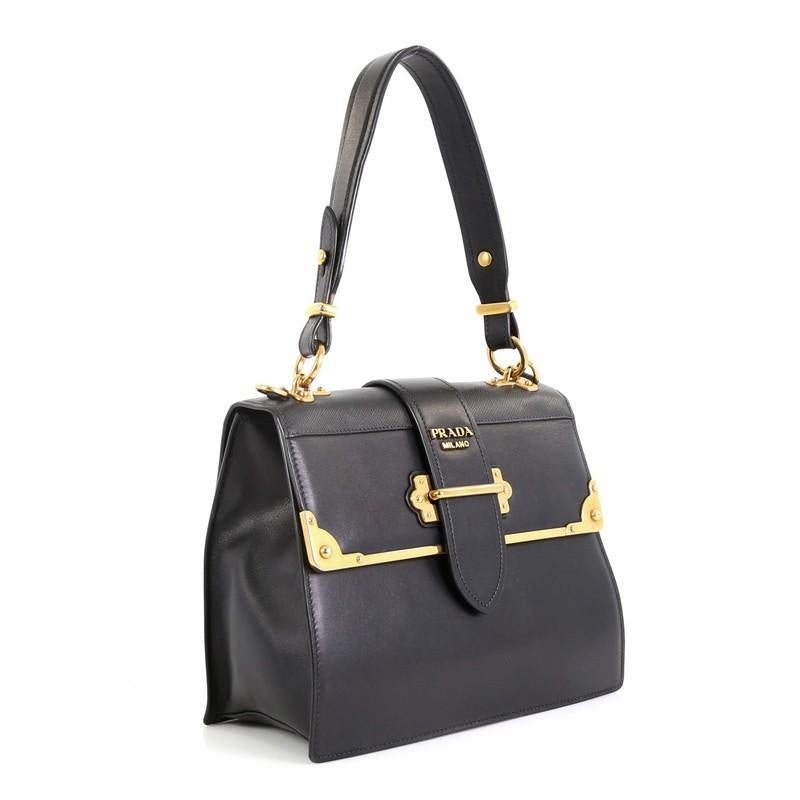 This Prada Cahier Convertible Shoulder Bag City Calf and Saffiano Leather Medium, crafted from black city calf and saffiano leather, features a flat leather strap, metal hardware trim, exterior back zip pocket, and gold-tone hardware. Its buckle