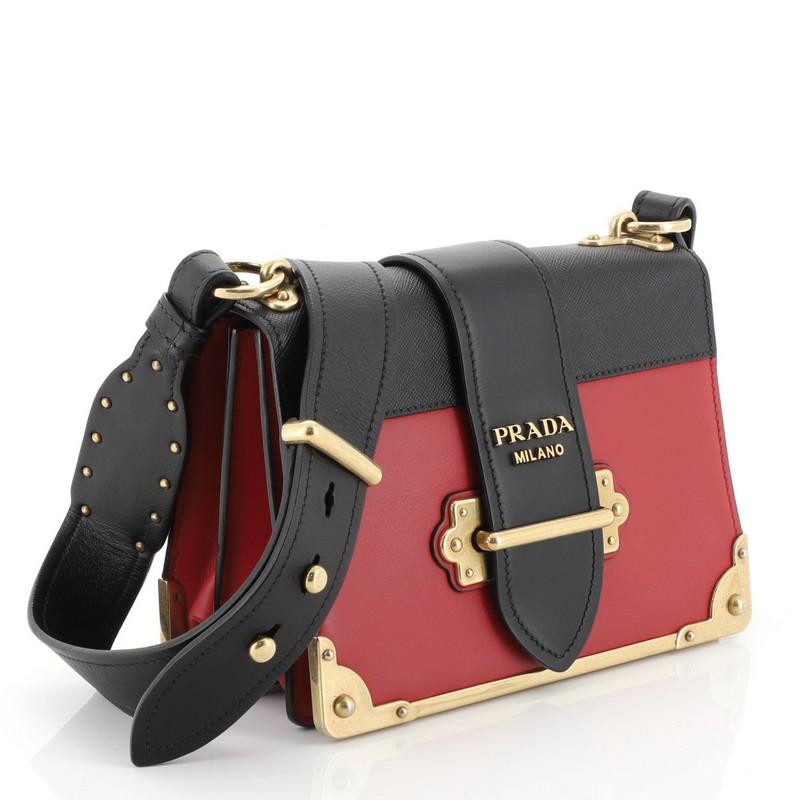 This Prada Cahier Crossbody Bag City Calf and Saffiano Leather Small, crafted from black and red leather, features adjustable leather strap, metal hardware trim, and aged gold-tone hardware. Its buckle closure opens to a black leather interior.