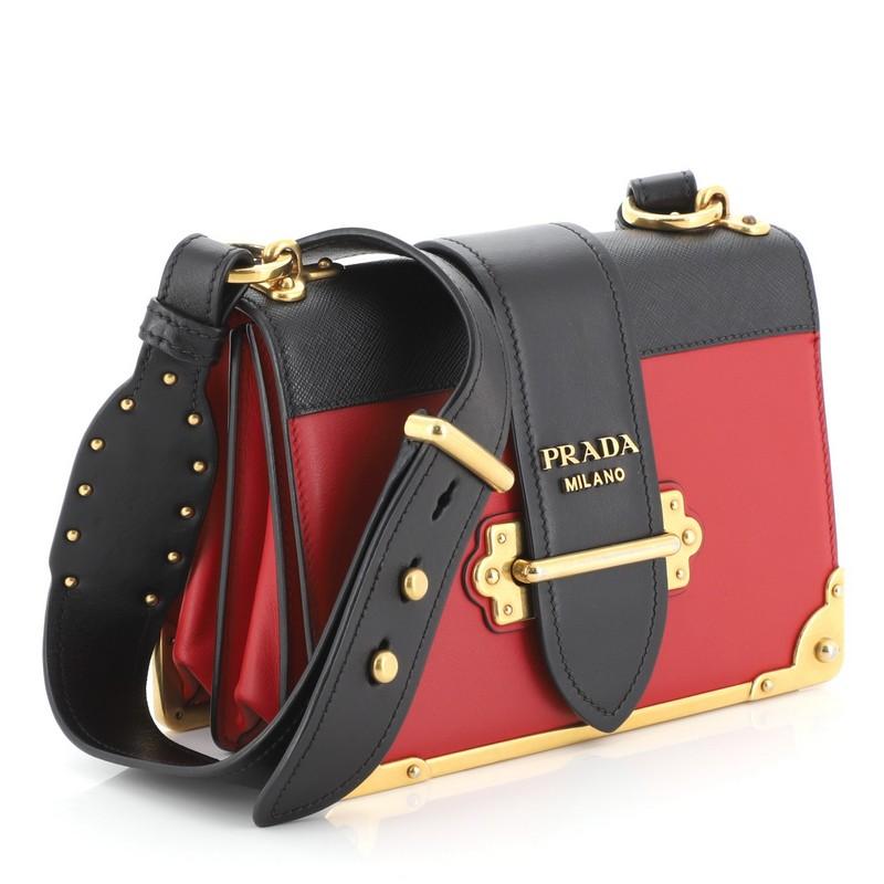 This Prada Cahier Crossbody Bag City Calf and Saffiano Leather Small, crafted from black and red leather, features adjustable leather strap, metal hardware trim, and aged gold-tone hardware. Its buckle closure opens to a black leather interior.