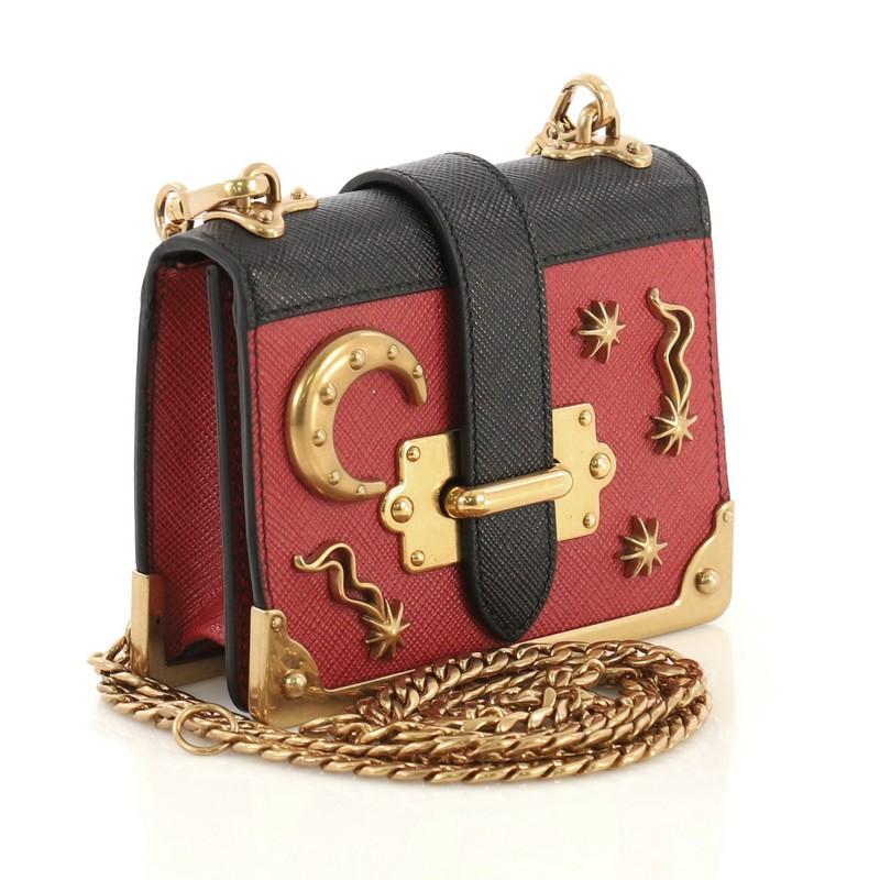 This Prada Cahier Crossbody Bag Embellished Leather Micro, crafted from black and red leather, features chain link strap, metal hardware trim, star and moon detailing, and aged gold-tone hardware. Its buckle closure opens to a black leather