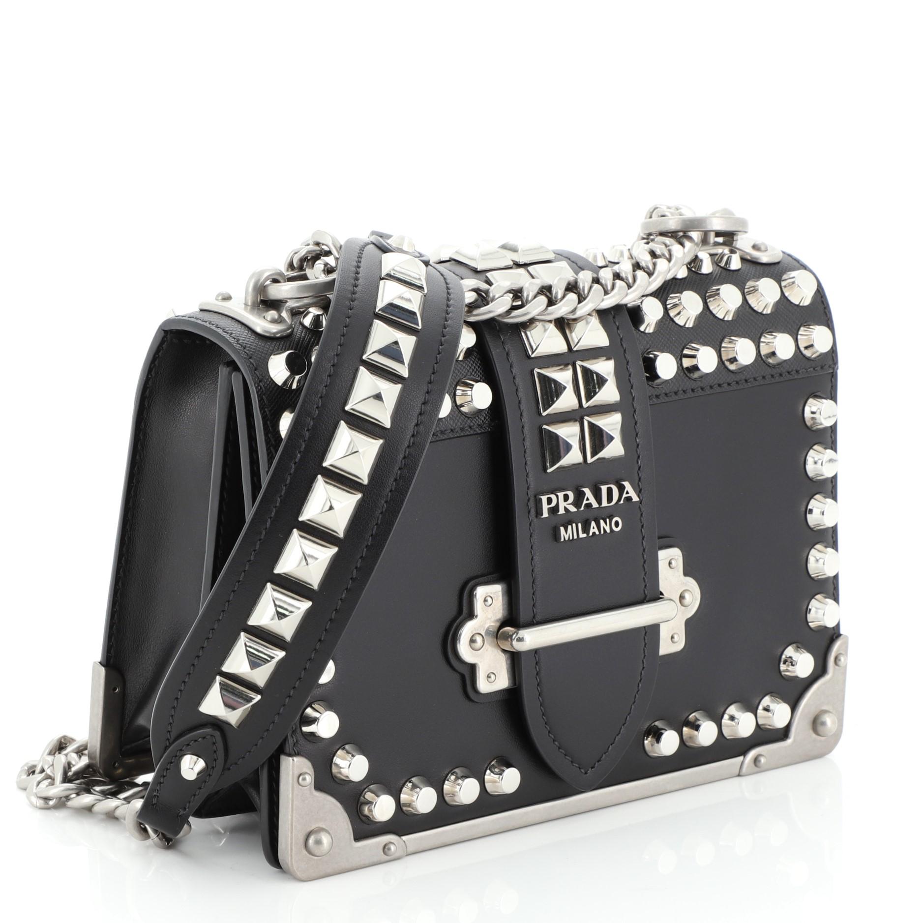 This Prada Cahier Crossbody Bag Studded City Calf with Saffiano Leather Small, crafted from black saffiano leather, features chain-link strap with leather shoulder pad, silver stud detailing, front flap with buckle closure and silver-tone hardware.