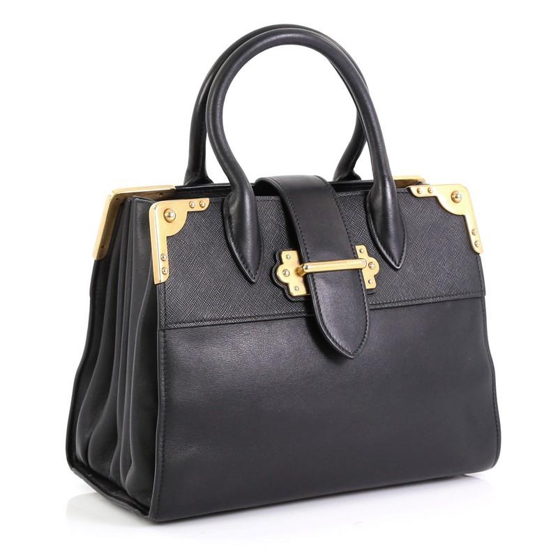 This Prada Cahier Tote City Calf and Saffiano Leather Large, crafted in black leather, features dual-rolled leather handles, metal hardware trim, and gold-tone hardware. Its flap tab with buckle closure opens to a black leather interior divided into