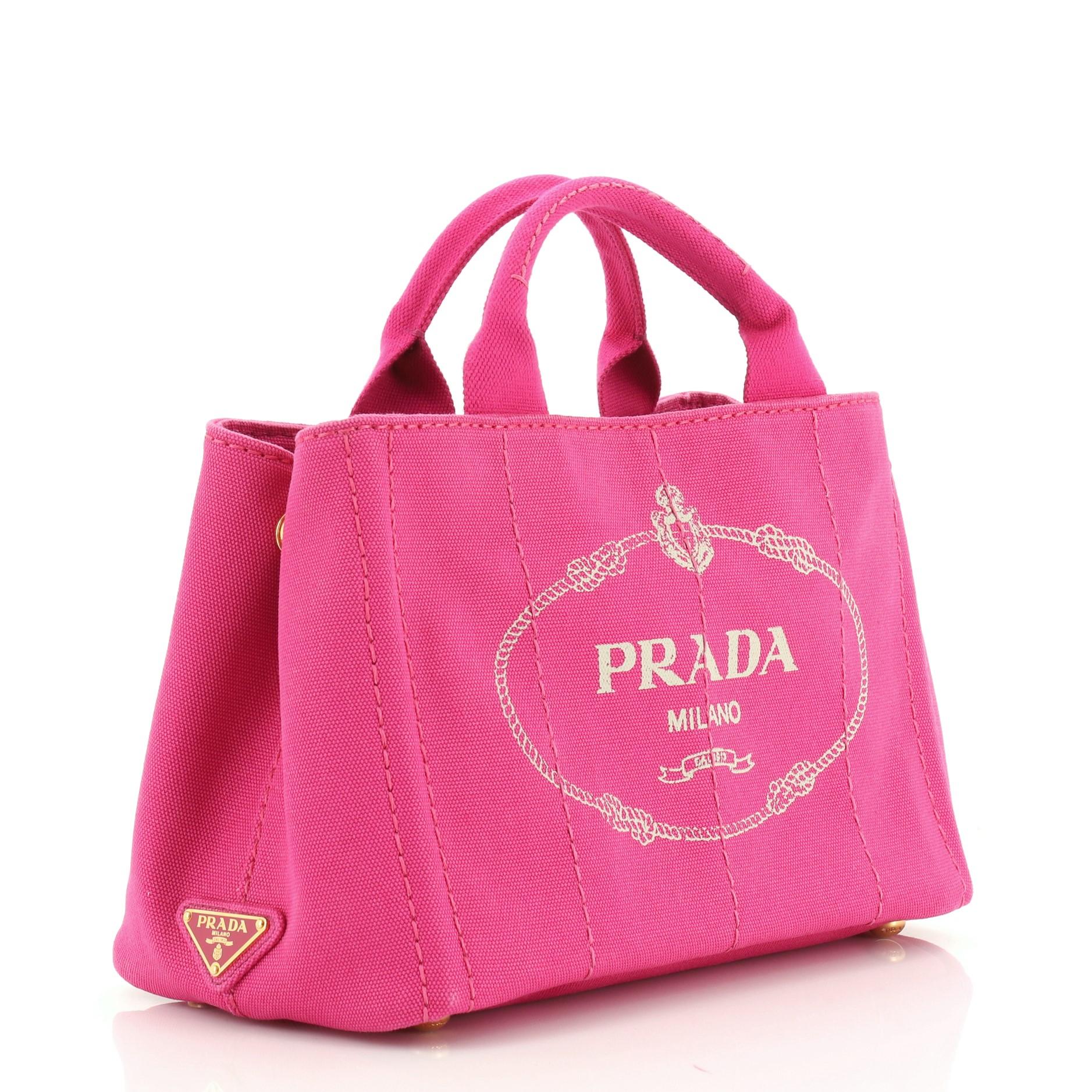 This Prada Canapa Convertible Tote Canvas Mini, crafted in pink canvas, features dual top handles, side snap buttons, protective base studs, and gold-tone hardware. It opens to a pink canvas interior with side zip and slip pockets. 

Estimated