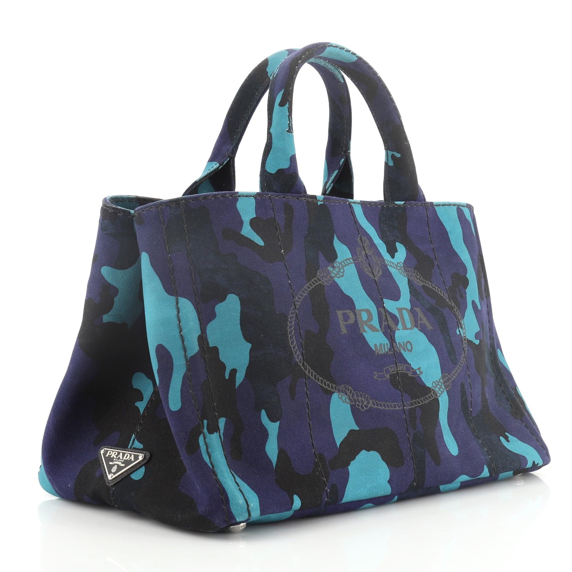 This Prada Canapa Convertible Tote Printed Canvas Medium, crafted from blue printed canvas, features dual-rolled handles, side snap buttons, and silver-tone hardware. It opens to a black fabric interior with zip and slip pockets. 

Estimated Retail