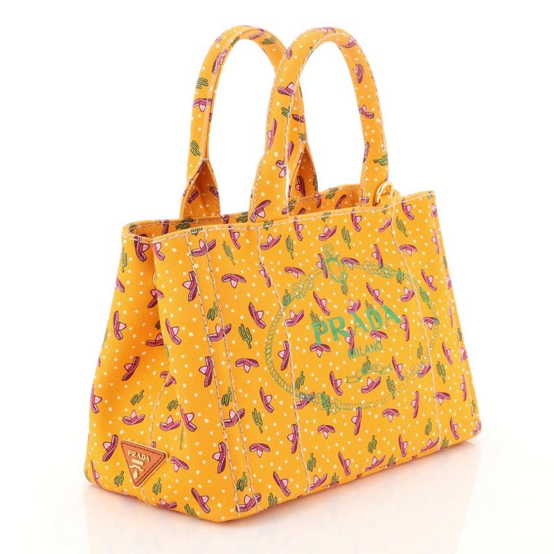 This Prada Canapa Convertible Tote Printed Canvas Mini, crafted from orange printed canvas, features dual rolled handles, side snap buttons, and gold-tone hardware. Its wide top opening showcases an orange printed canvas interior with zip and slip