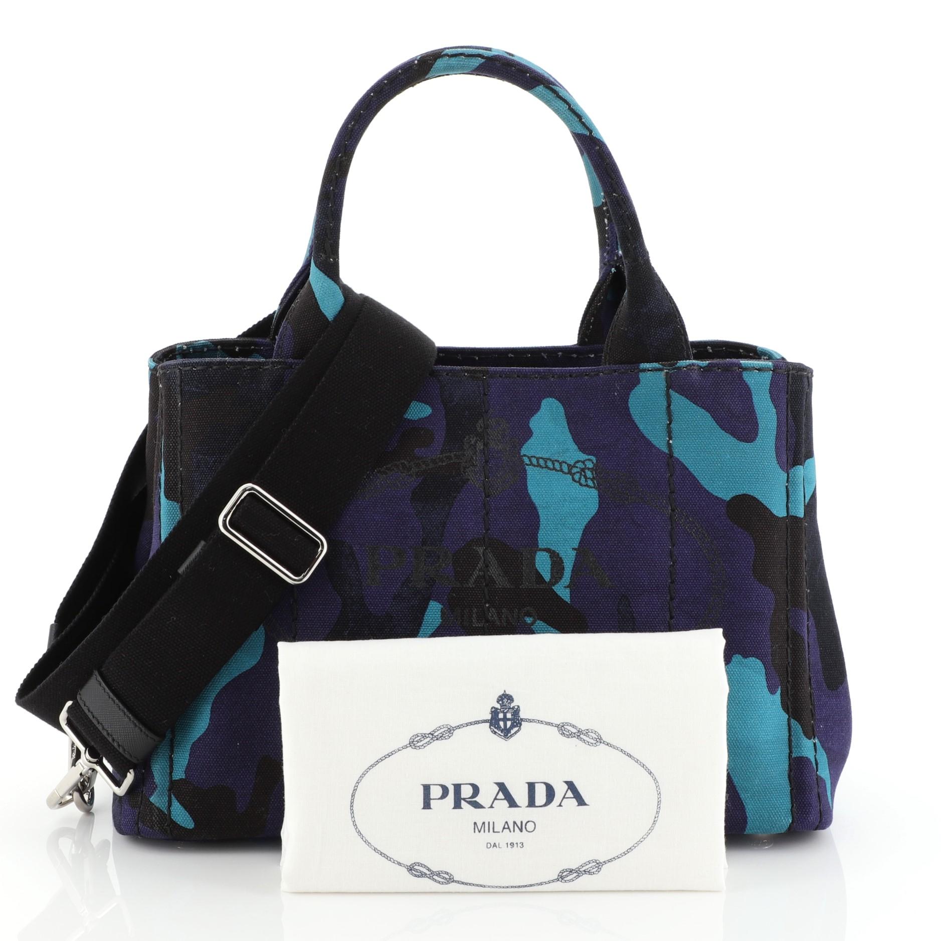 This Prada Canapa Convertible Tote Printed Canvas Mini, crafted from blue and purple printed canvas, features dual rolled handles, side snap buttons, and silver-tone hardware. It opens to a black canvas interior with zip and slip pockets.