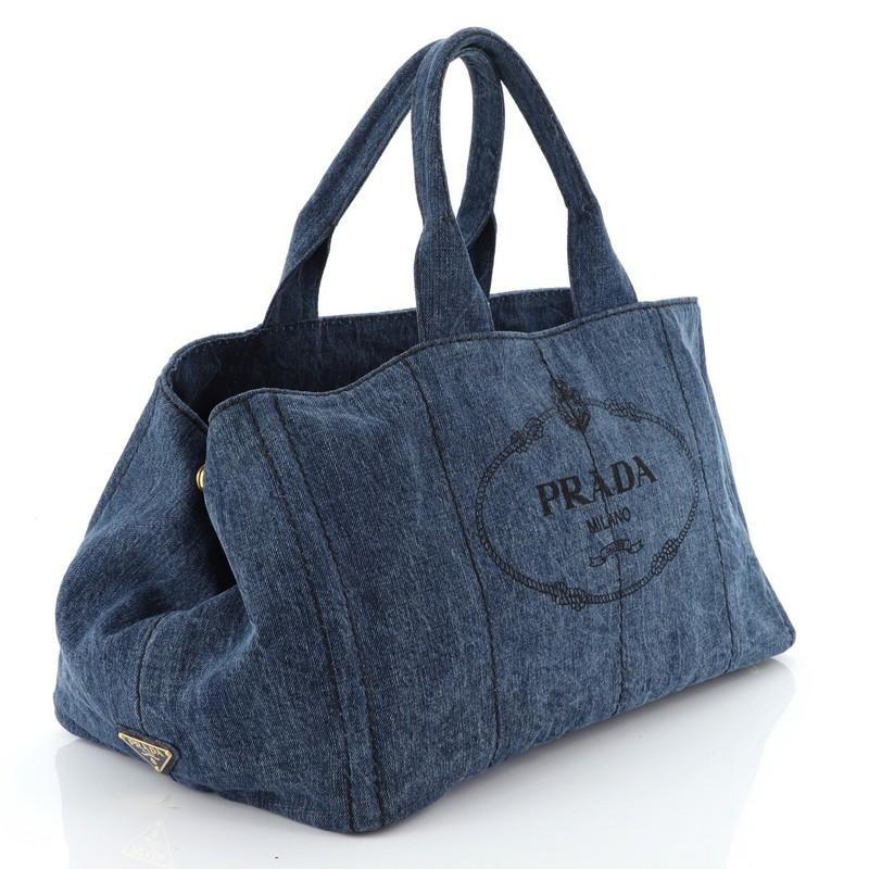 This Prada Canapa Tote Denim Large, crafted in blue denim, features dual rolled handles, Prada logo at the center, side snap buttons, protective base studs and gold-tone hardware. It opens to a blue denim interior with side zip and slip pockets.