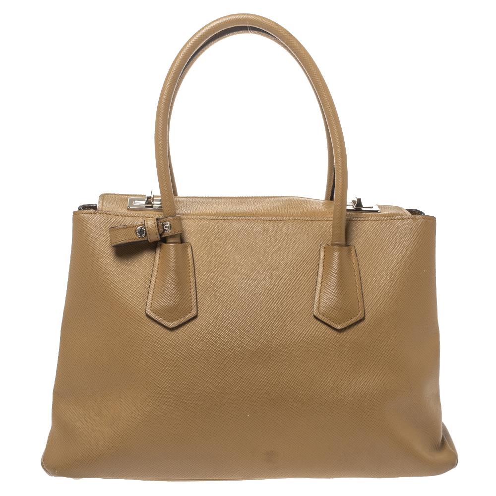 Stay on top of your fashion game with this Twin tote from Prada! Crafted from Saffiano leather, the tote has a caramel brown hue with two rolled handles, a leather tag, and protective metal feet. The insides are leather-lined and sized to dutifully